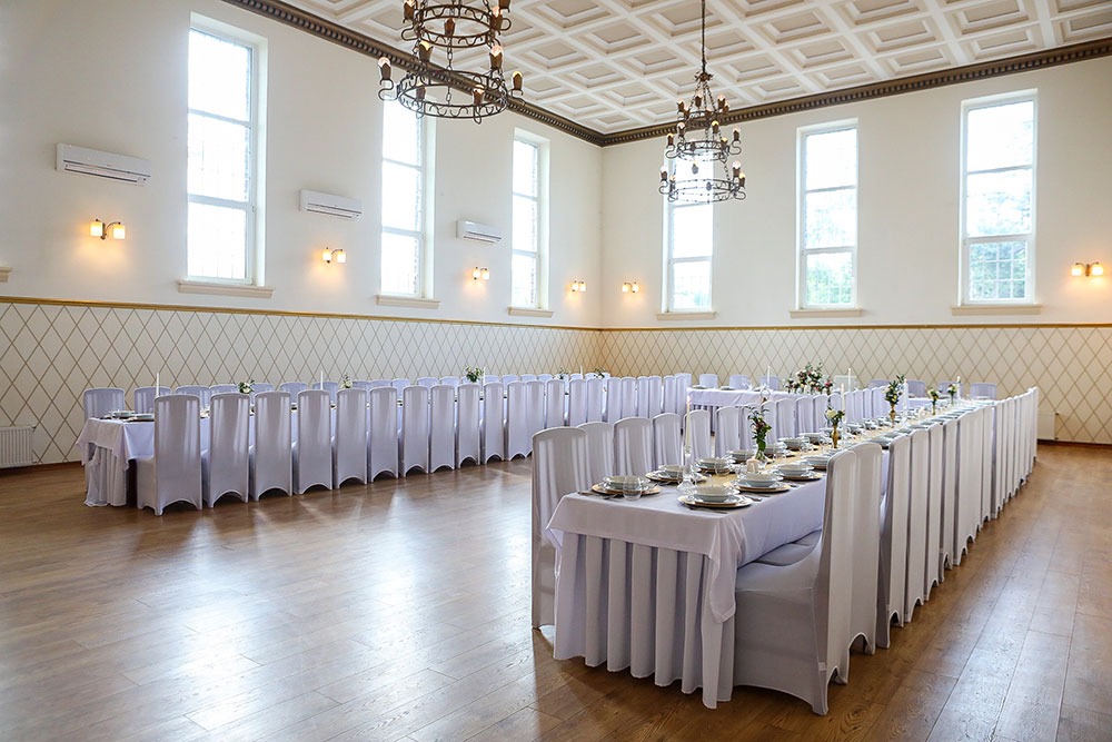 Sala bankietowa z długimi stołami nakrytymi białymi obrusami i krzesłami w pokrowcach, dekoracje kwiatowe na stołach, parkiet, wysokie okna z klimatyzacją, żyrandole w stylu retro.