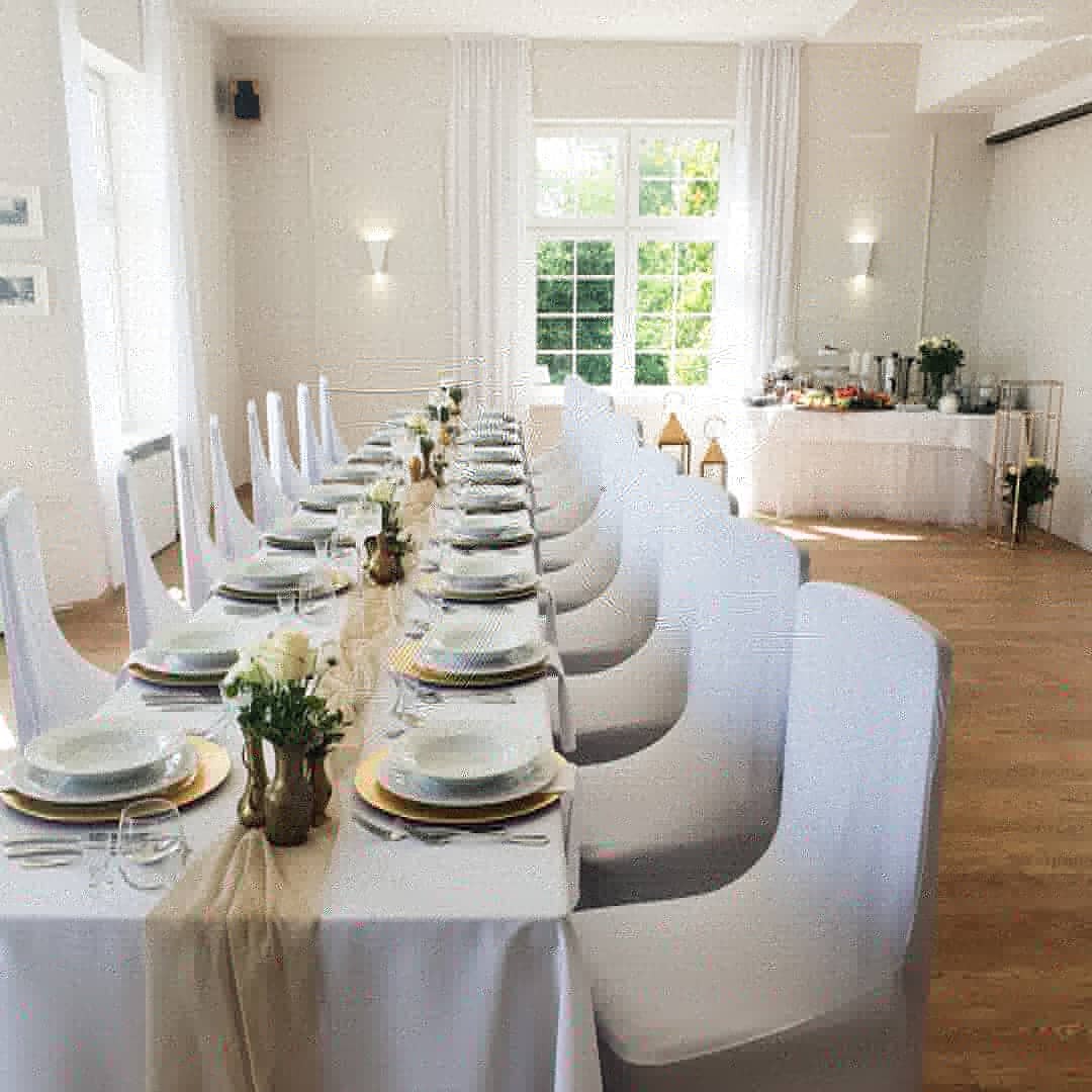 Długi stół udekorowany białym obrusem, bieżnikiem w kolorze beżowym, złotymi podkładkami pod talerze, białymi talerzami, sztućcami i szklankami, z białymi różami w złotych wazonach, krzesła...