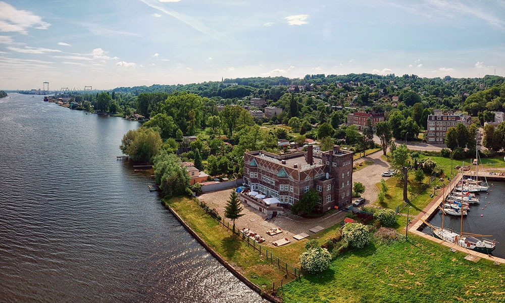 Widok z lotu ptaka na ceglany budynek z patio nad rzeką, z miejscami do siedzenia na zewnątrz i zacumowanymi łodziami w pobliżu, otoczony zielenią, z widocznymi budynkami w tle.
