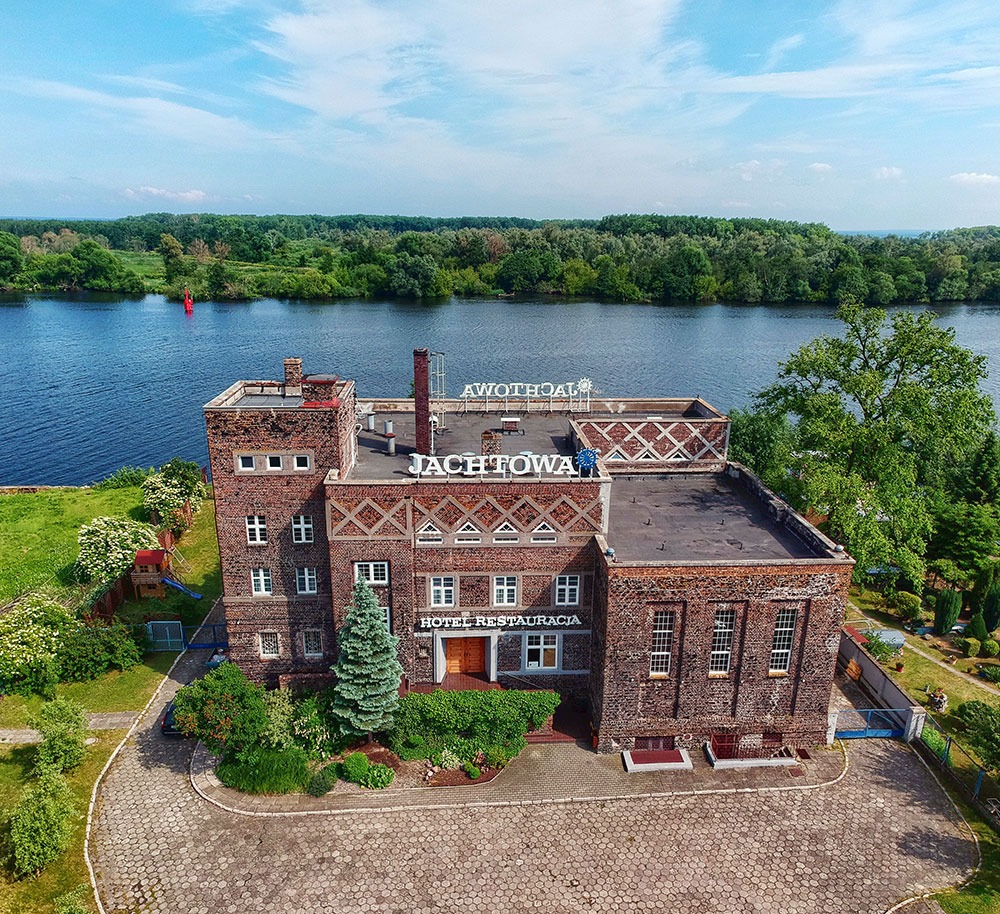 Widok z lotu ptaka na budynek hotelu i restauracji Jachtowa, położony nad rzeką, z widocznym ogrodem i parkingiem.