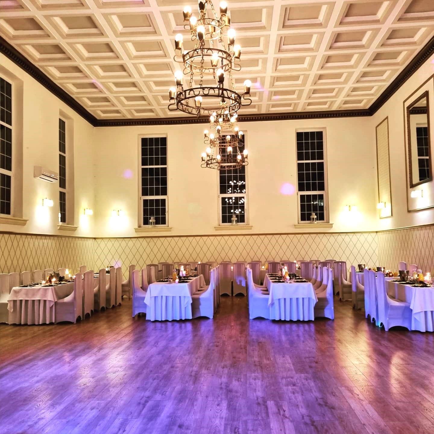 Sala bankietowa z nakrytymi stołami, krzesła w pokrowcach, oświetlenie świecami i żyrandolami, parkiet z fioletowym refleksem.