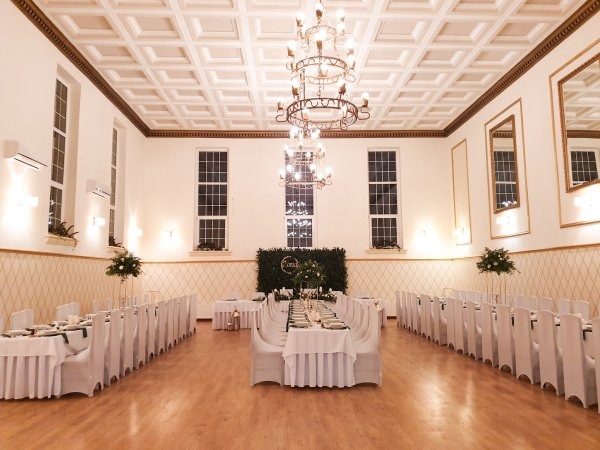 Uroczysta sala bankietowa w Szczecinie, przygotowana na przyjęcie, z długimi stołami nakrytymi białymi obrusami i krzesłami w pokrowcach, dekoracje kwiatowe i eleganckie żyrandole.