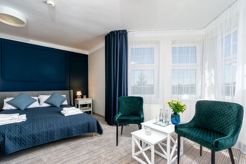 Nowoczesny pokój hotelowy z ciemnoniebieską ścianą, szarą tapicerowaną ramą łóżka, ciemnozielonymi krzesłami i białymi stolikami kawowymi, bukietem żółtych kwiatów i butelkami wody mineralnej