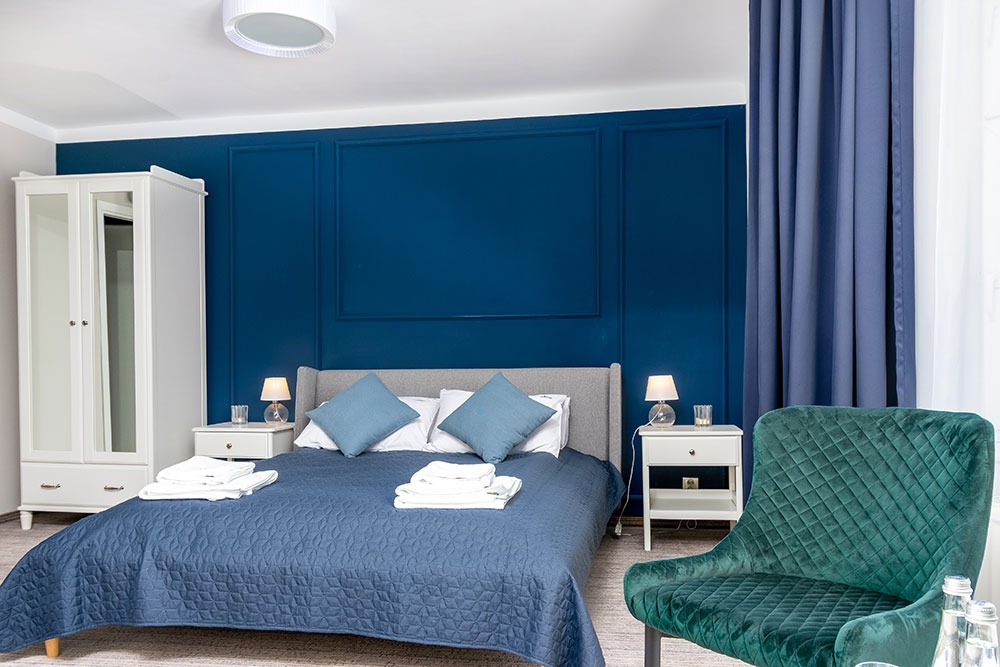 Nowoczesny pokój hotelowy z ciemnoniebieską ścianą z dekoracyjnymi panelami, szarą tapicerowaną ramą łóżka, niebieską narzutą, białymi szafkami nocnymi z lampkami i zielonym aksamitnym krzesłem...