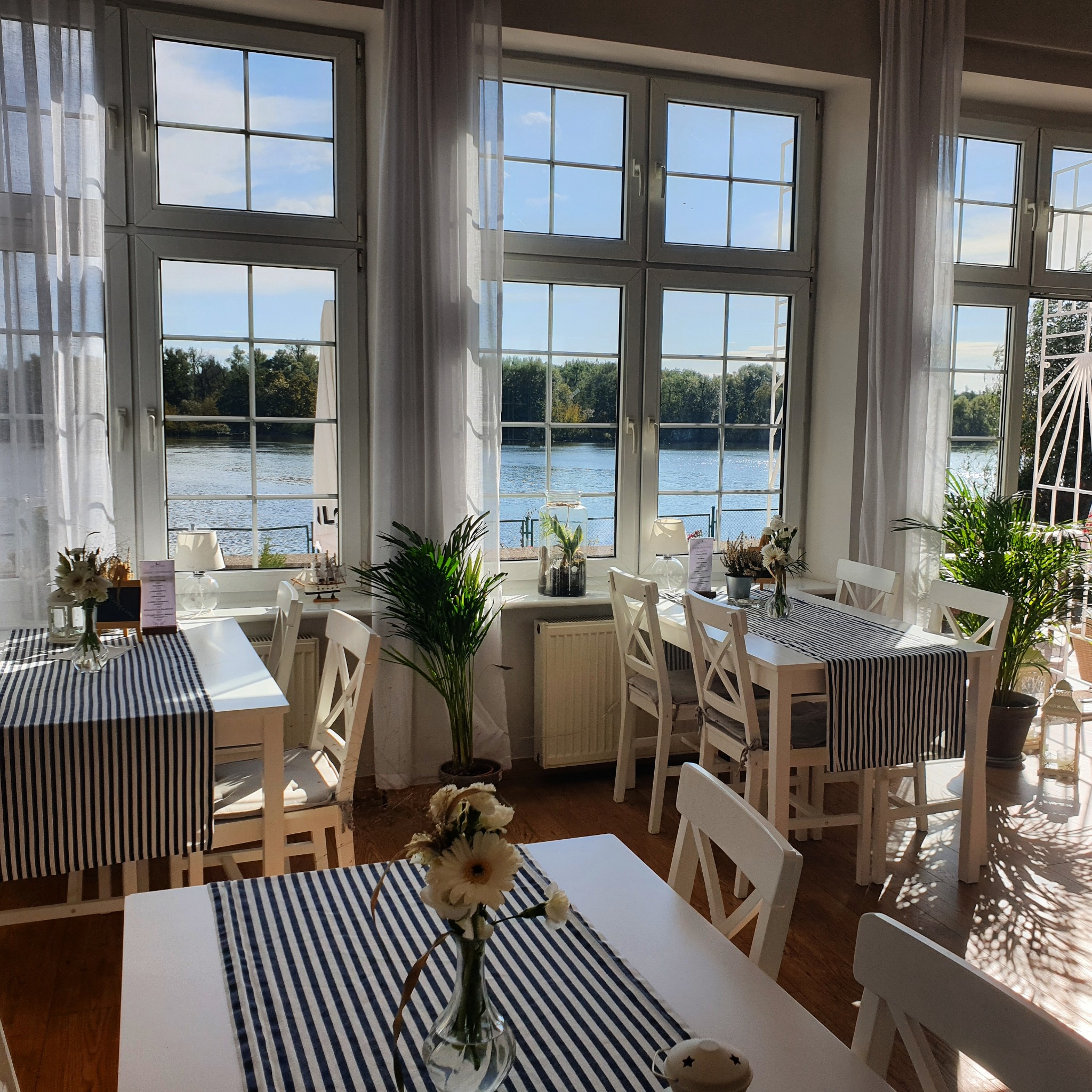 Jasne wnętrze restauracji z białymi stołami nakrytymi w paski, widok na rzekę przez duże okna z białymi firankami, dekoracje kwiatowe i roślinne.