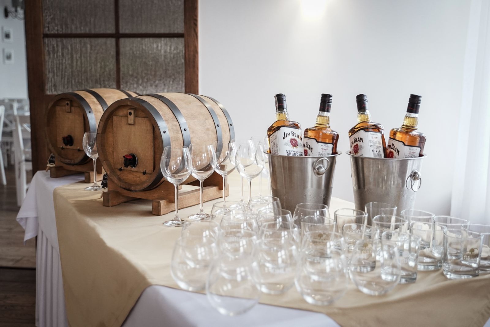Rustykalny bar z dwoma drewnianymi beczkami, czterema butelkami Jim Beam w metalowych wiaderkach z lodem oraz różnorodnymi szklankami i kieliszkami na beżowym obrusie.