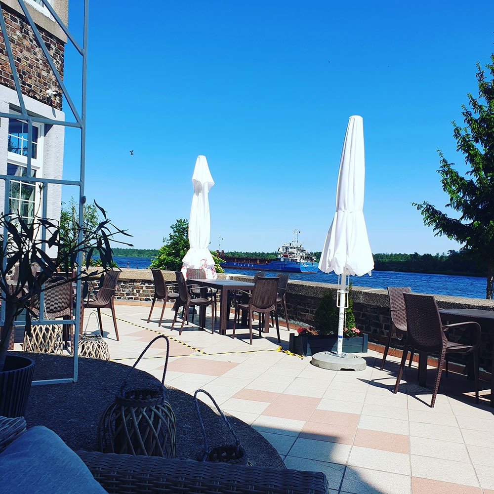 Taras restauracyjny z widokiem na rzekę i przepływający statek transportowy, białe parasole złożone, brązowe krzesła i stoły na tle błękitnego nieba.
