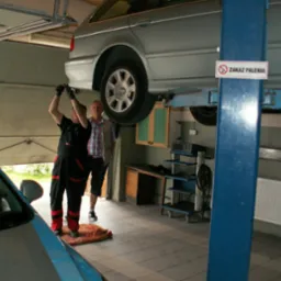 Srebrne kombi na podnośniku w warsztacie; mechanik w czarnym kombinezonie z czerwonymi wstawkami przy pojeździe, drugi mężczyzna w koszuli w kratę obserwuje, w tle biurko, półki i znak zakazu palenia.