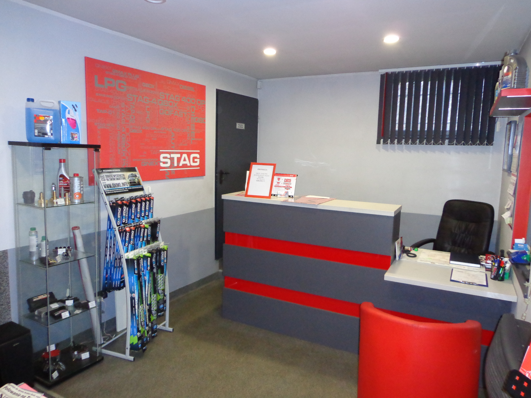 Wnętrze biura warsztatu samochodowego z recepcją w szaro-czerwonych barwach, ekspozytorem z płynami i akcesoriami samochodowymi, oraz plakatem informacyjnym marki Stag.