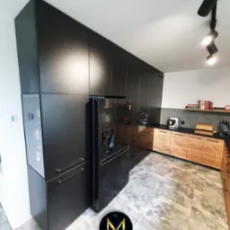 Nowoczesna kuchnia z czarnymi szafkami sięgającymi sufitu, drewnianymi frontami dolnych szafek, czarnym blatem i lodówką w zabudowie, oświetlona reflektorami sufitowymi.