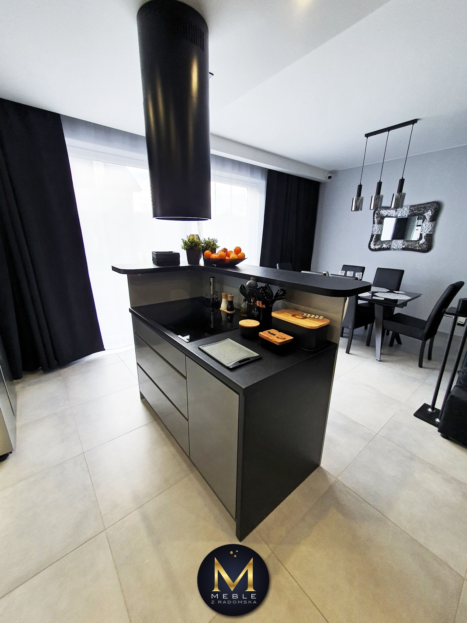 Nowoczesna wyspa kuchenna z czarnym blatem i szarymi frontami, widok na stół jadalny i dekoracyjny lustro, czarny okap cylindryczny, logo firmy w prawym dolnym rogu.