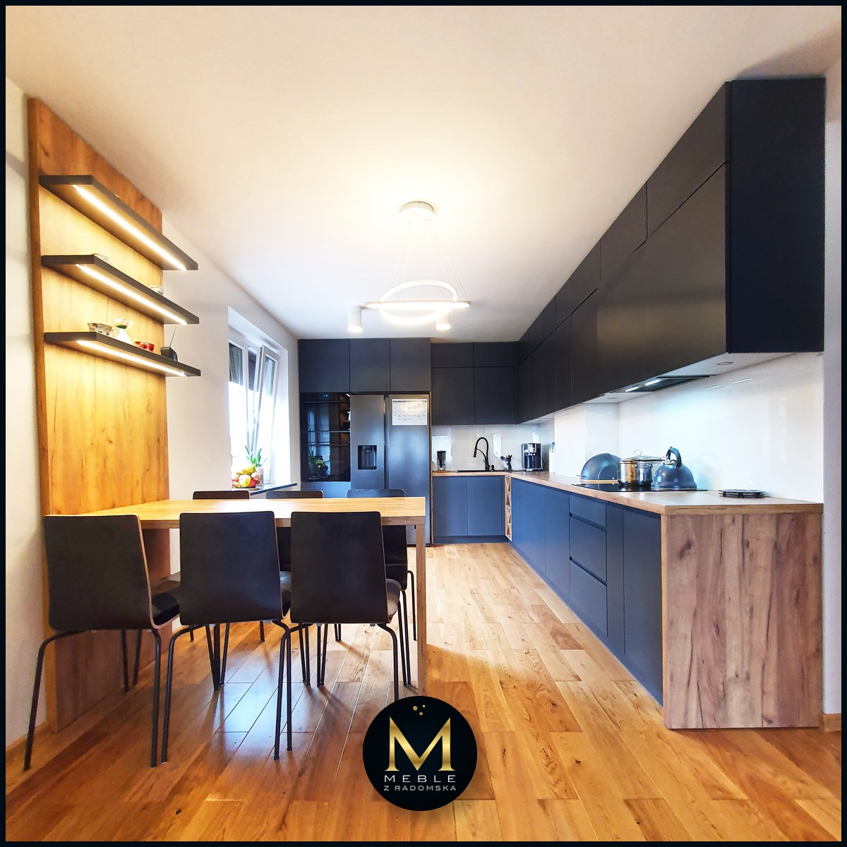 Nowoczesna kuchnia na wymiar z ciemnymi frontami i drewnianym blatem, oświetlenie LED na półkach i pod szafkami, drewniana podłoga, stół z krzesłami w tle.