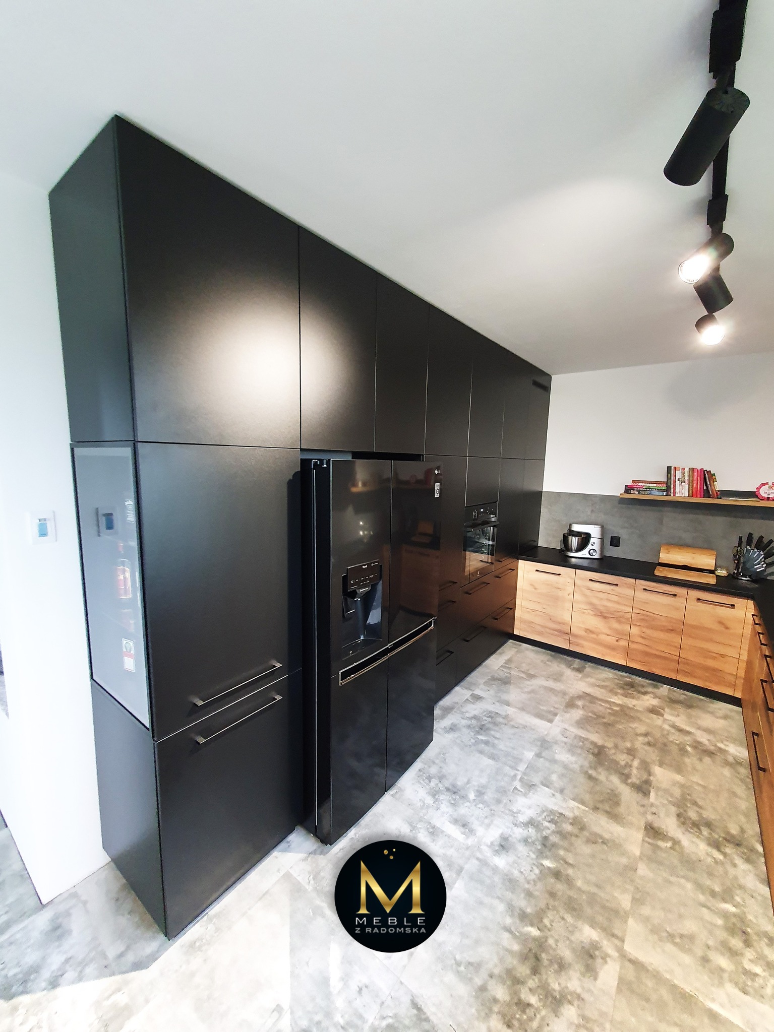 Nowoczesna kuchnia z czarnymi szafkami sięgającymi sufitu, drewnianymi frontami dolnych szafek, czarnym blatem i lodówką w zabudowie, oświetlona reflektorami sufitowymi.