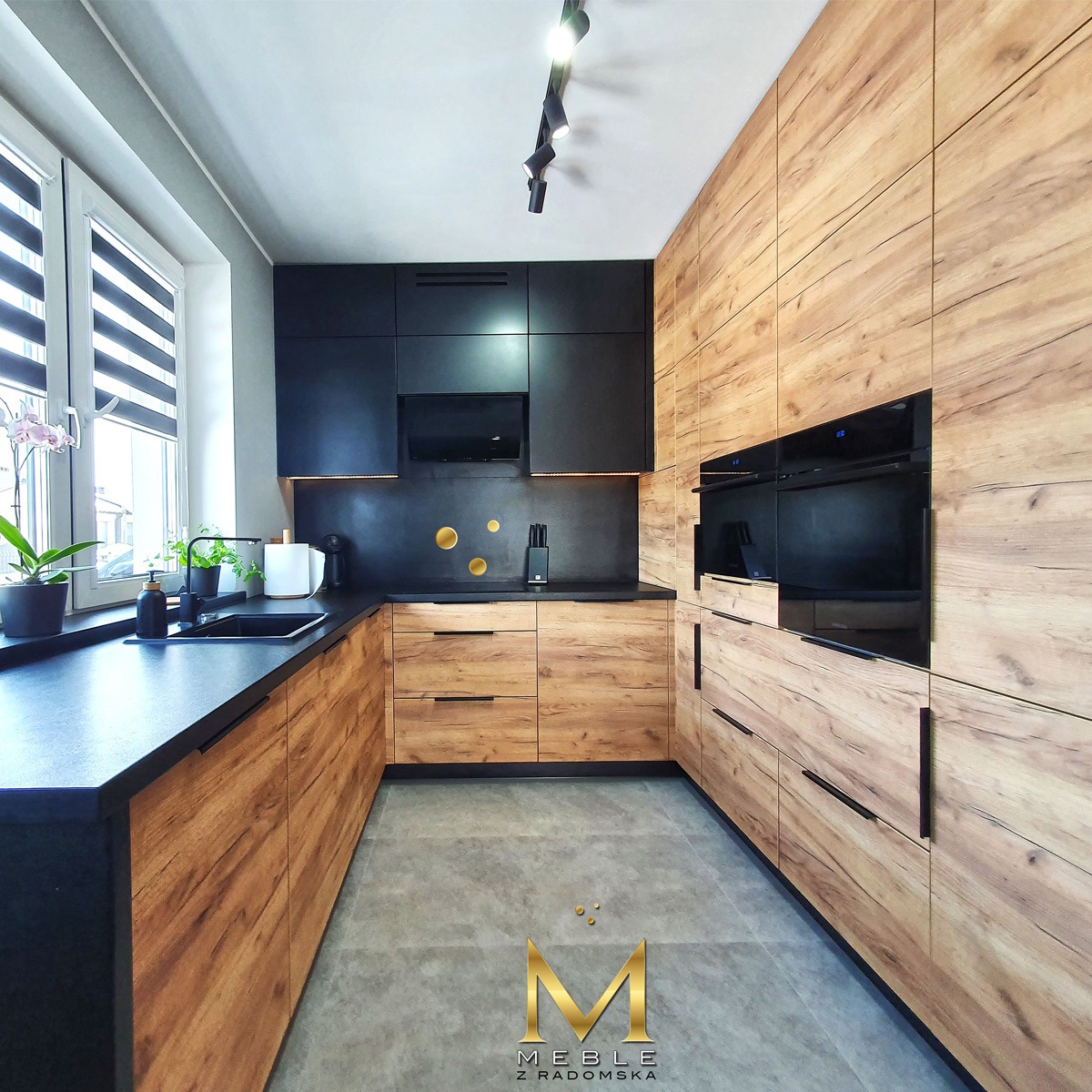 Nowoczesna kuchnia w odcieniach drewna i czerni, z blatem z ciemnego kamienia, minimalistycznym oświetleniem i elementami dekoracyjnymi w postaci złotych kropek na ścianie. Na blacie widoczne...