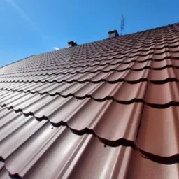 Widok z dołu na nowy, brązowy dach z blachodachówki modułowej, z widocznymi kominami i anteną na tle błękitnego nieba.