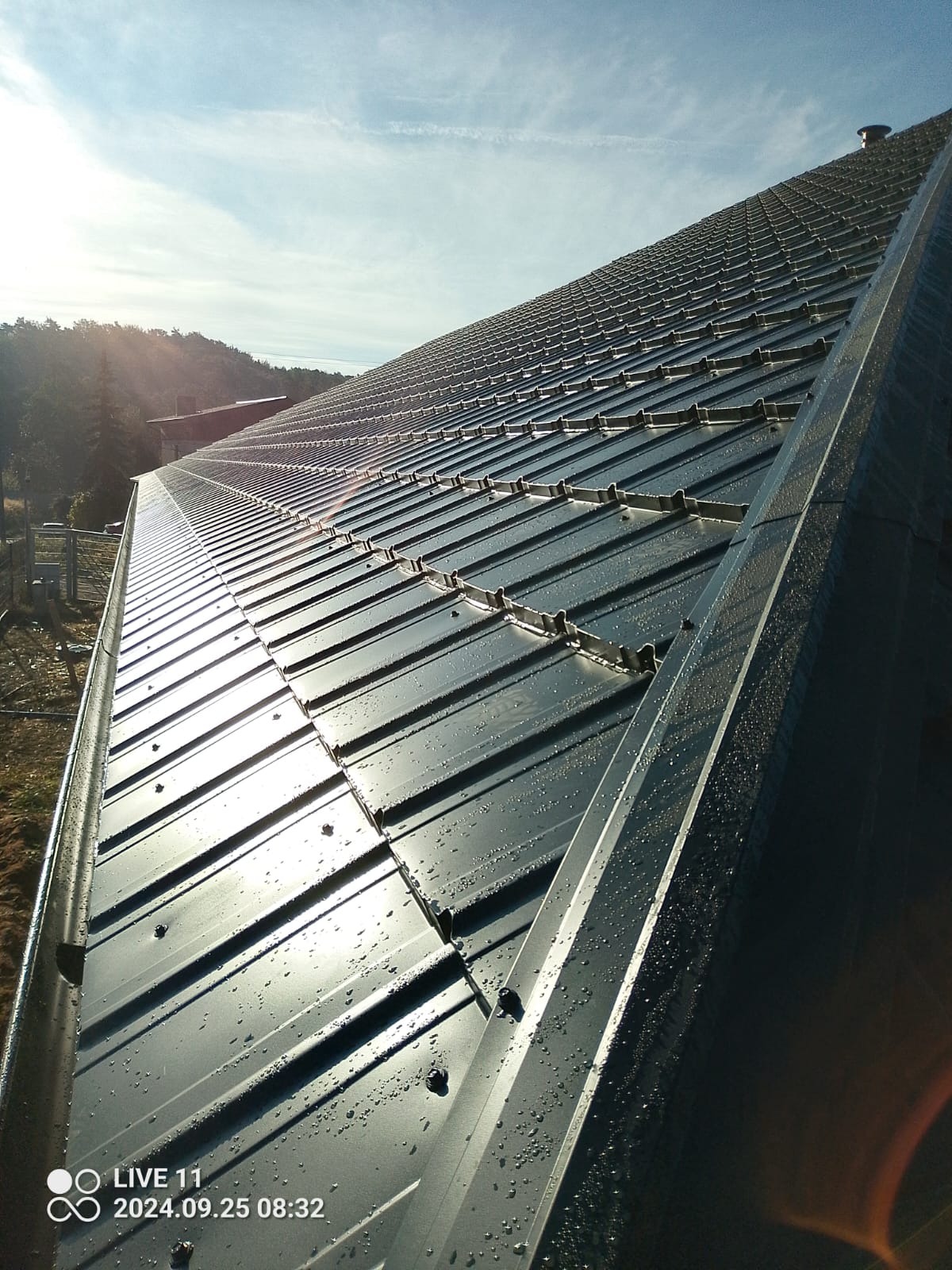 Nowy, błyszczący dach z blachy na skośnej konstrukcji, pokryty kroplami rosy w porannym słońcu, z widoczną obróbką blacharską i fragmentem rynny.