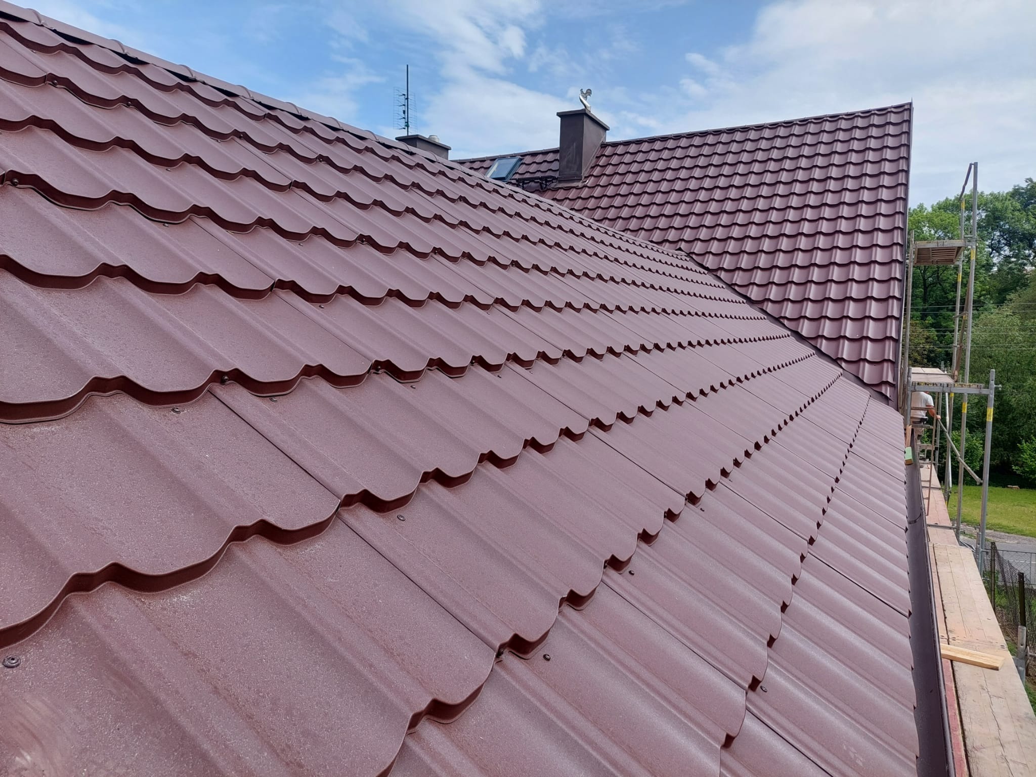 Czerwono-brązowe metalowe dachówki na skośnym dachu, widoczne kominy i antena, częściowo widoczne rusztowanie w tle, błękitne niebo z chmurami.