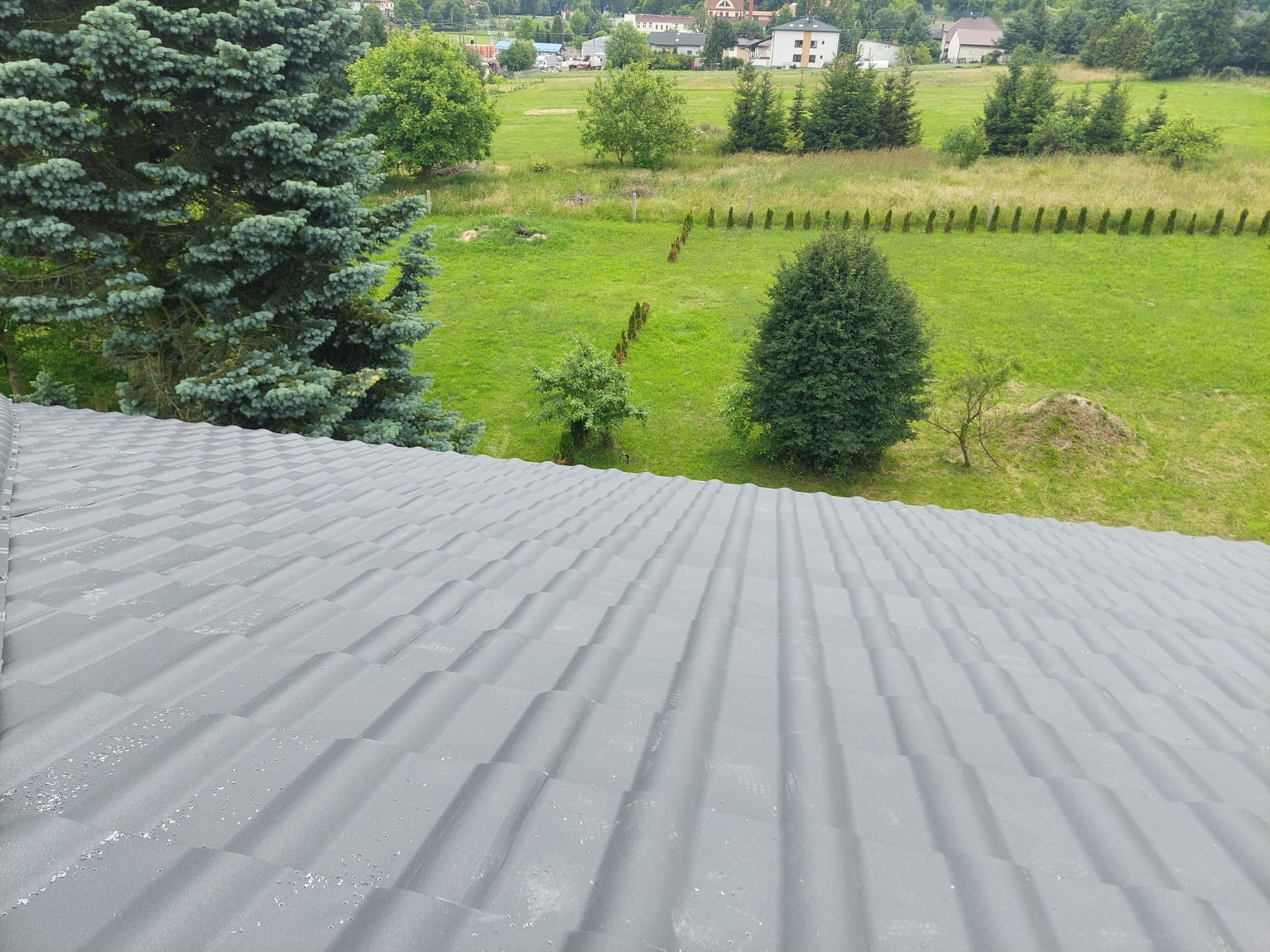 Szary dach z blachy trapezowej, widok z góry na tle zielonego krajobrazu z drzewami i zabudowaniami w oddali.