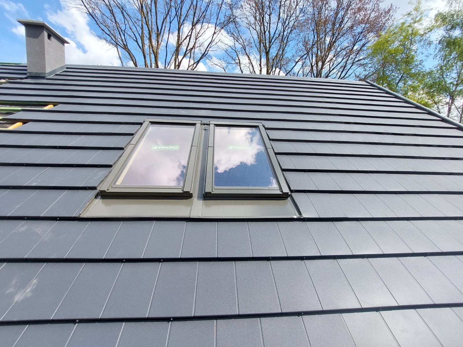 Szary dach z dachówki ceramicznej z dwoma oknami dachowymi marki Fakro, odbijającymi błękitne niebo z chmurami.