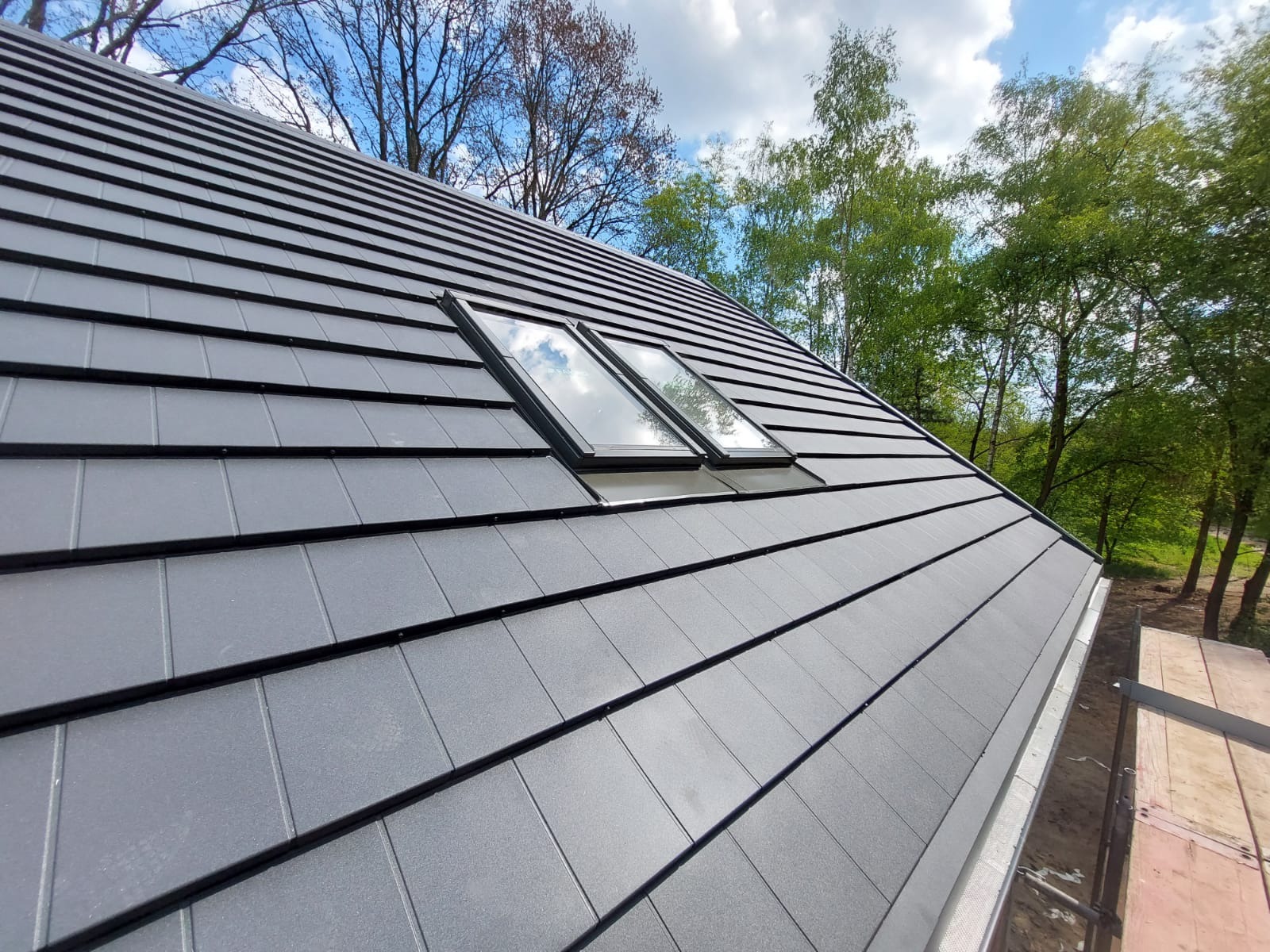Nowoczesne pokrycie dachu z szarej dachówki z dwoma oknami dachowymi, widoczne drzewa w tle.