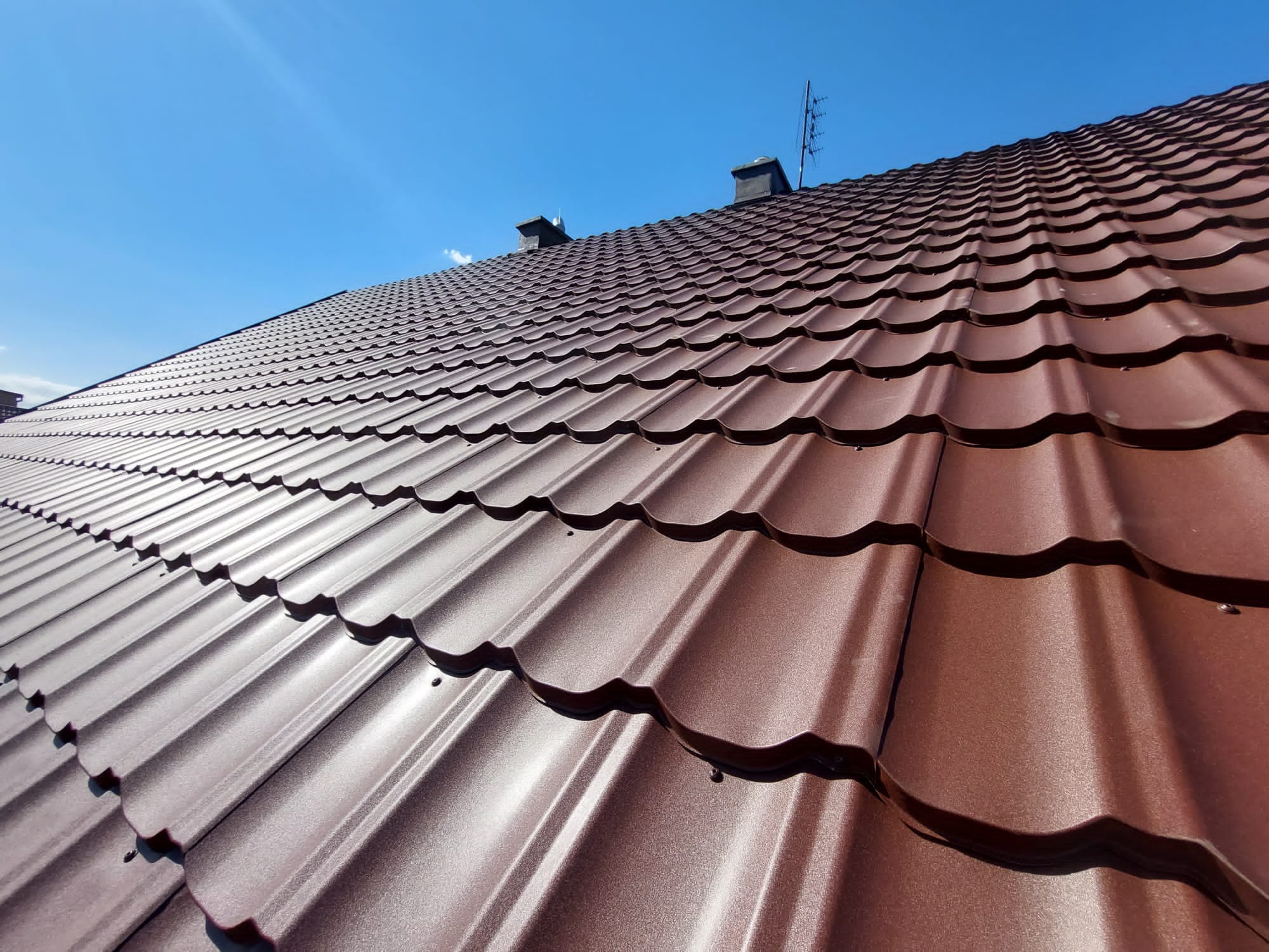 Widok z dołu na nowy, brązowy dach z blachodachówki modułowej, z widocznymi kominami i anteną na tle błękitnego nieba.