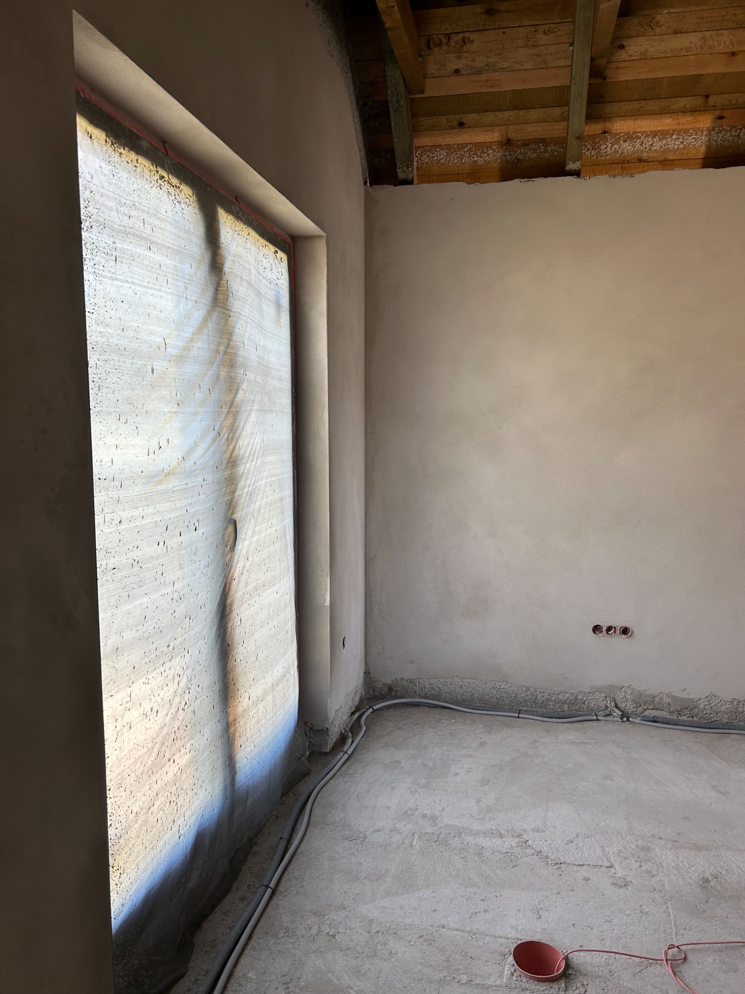 Świeżo położone tynki maszynowe na ścianach wewnętrznych budynku w trakcie budowy, widoczne kable elektryczne przy podłodze i drewniana konstrukcja dachu.