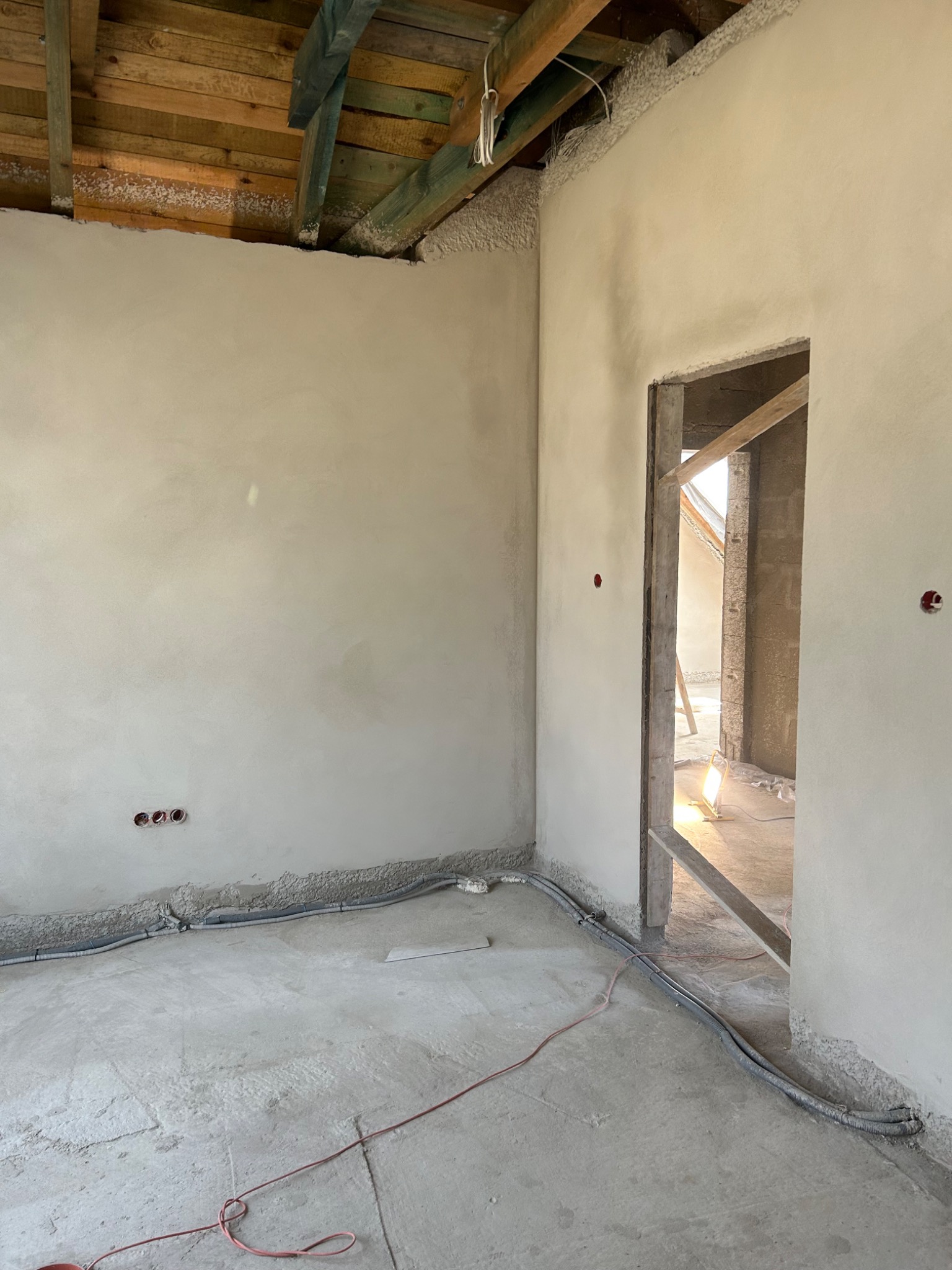 Wnętrze w trakcie prac wykończeniowych: ściany pokryte świeżym, jasnym tynkiem, widoczne przewody elektryczne przy podłodze, surowy drewniany strop i widok na sąsiednie pomieszczenie przez...