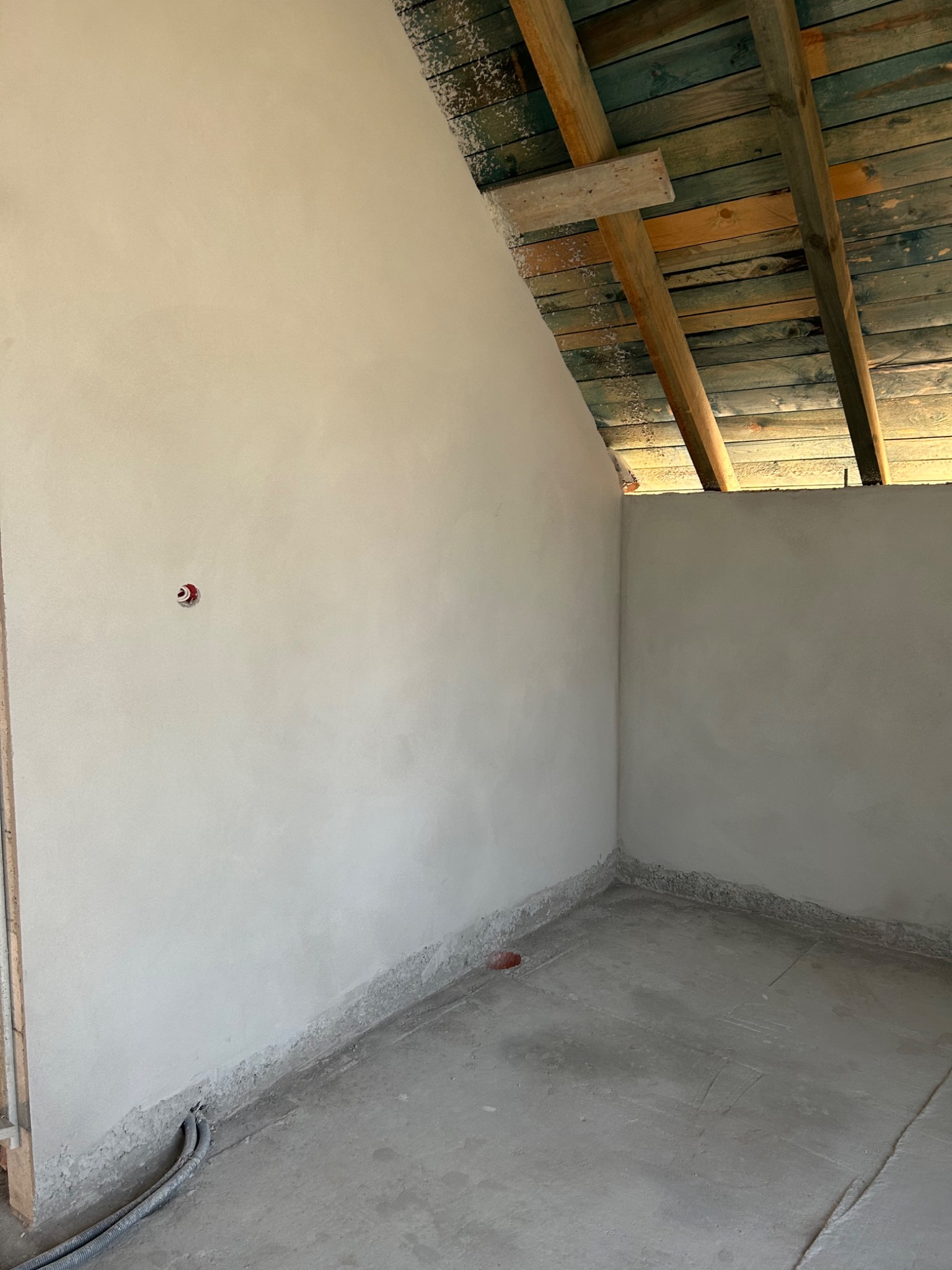 Świeżo otynkowane ściany poddasza z widoczną instalacją elektryczną i surowym drewnianym stropem, ukazujące etap prac wykończeniowych.