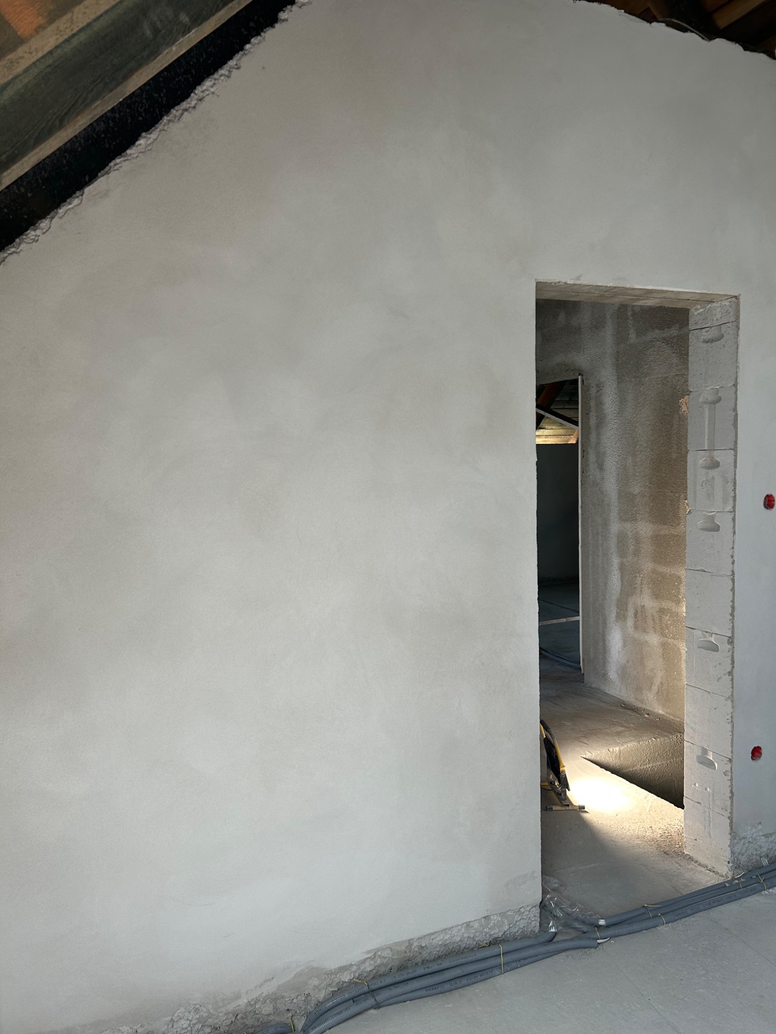 Świeżo położony, jasnoszary tynk na ścianie w surowym wnętrzu z widocznym otworem drzwiowym z betonu komórkowego, przez który widać dalszą część pomieszczenia. Na podłodze szare rury instalacyjne.