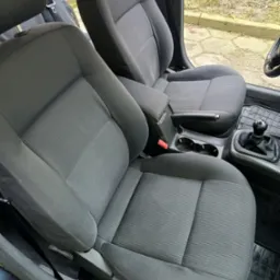 Wnętrze samochodu z widocznymi szarymi fotelami z tkaniny, czarną gałką zmiany biegów i gumowymi dywanikami