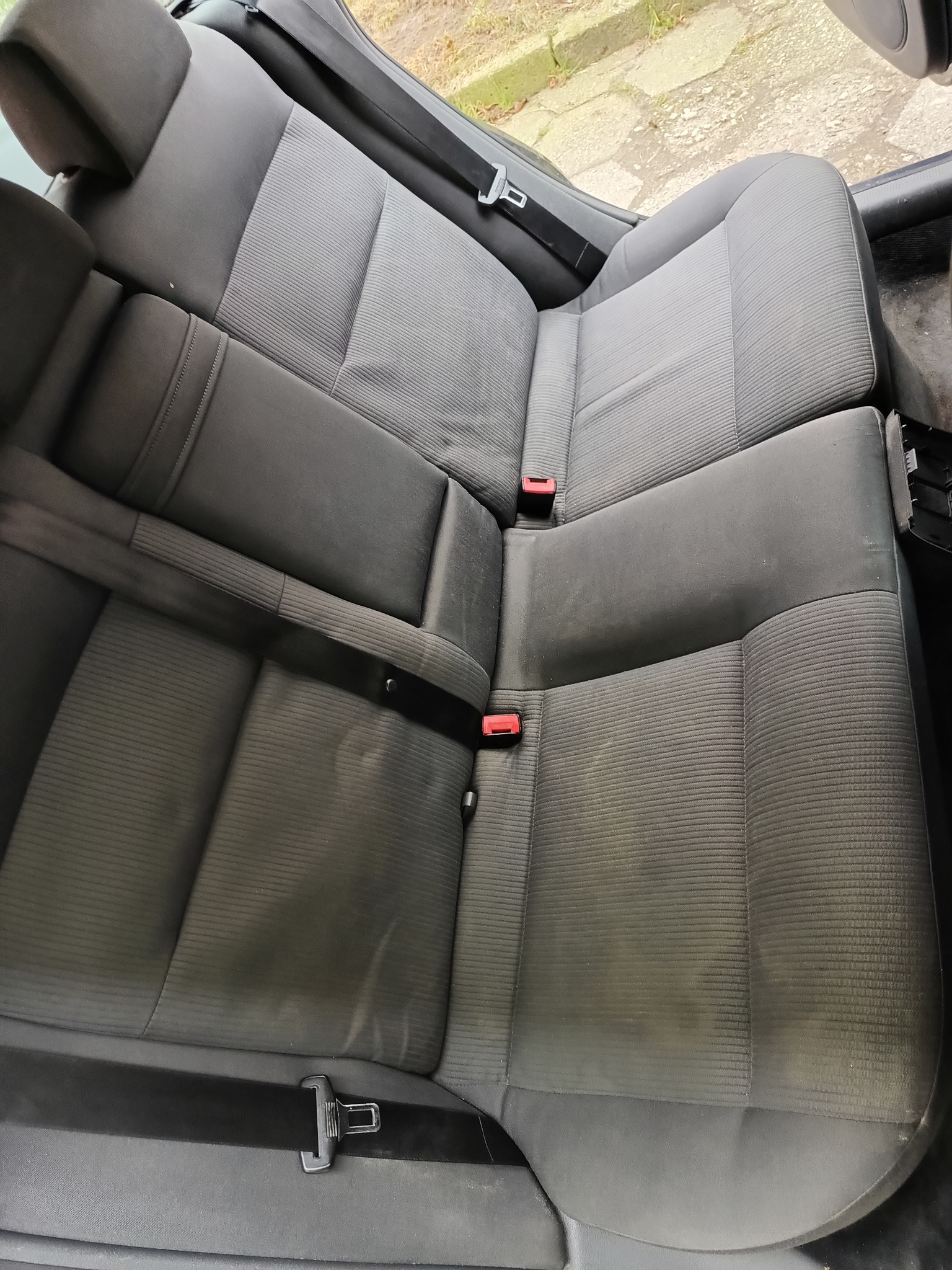 Szare, materiałowe siedzenie samochodowe z widocznymi plamami i zapiętymi pasami bezpieczeństwa.