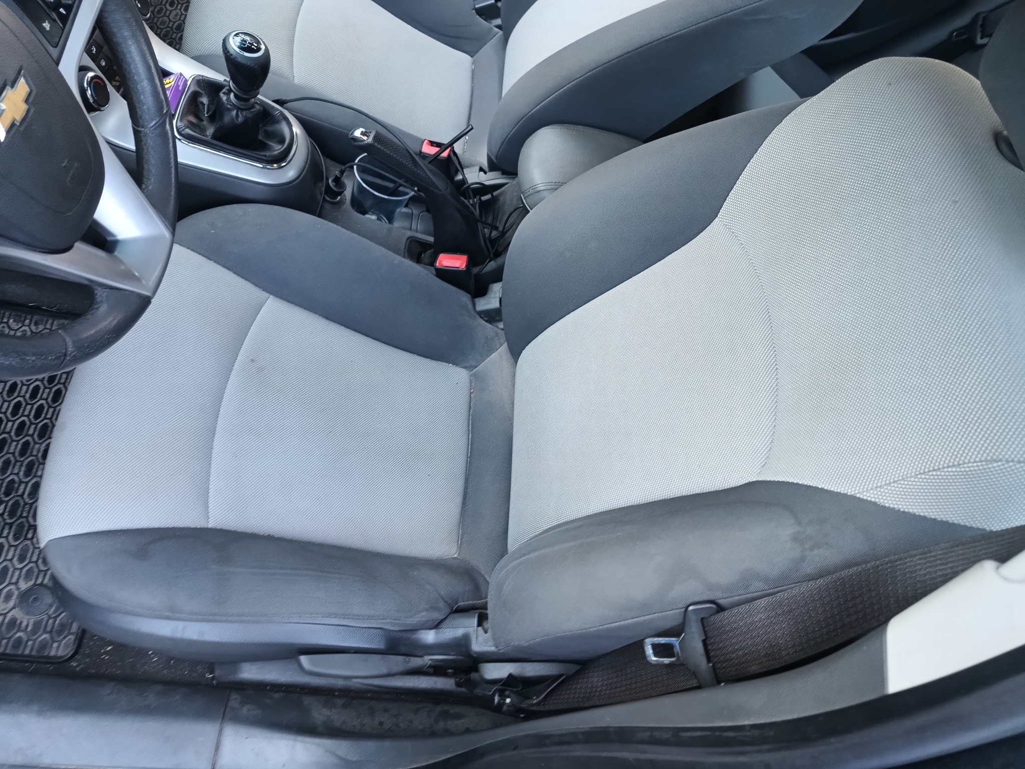 Wnętrze samochodu z widocznymi jasnoszarymi fotelami z ciemnymi elementami. Na fotelu kierowcy widoczne ślady użytkowania. Widoczna kierownica, dźwignia zmiany biegów i konsola środkowa.