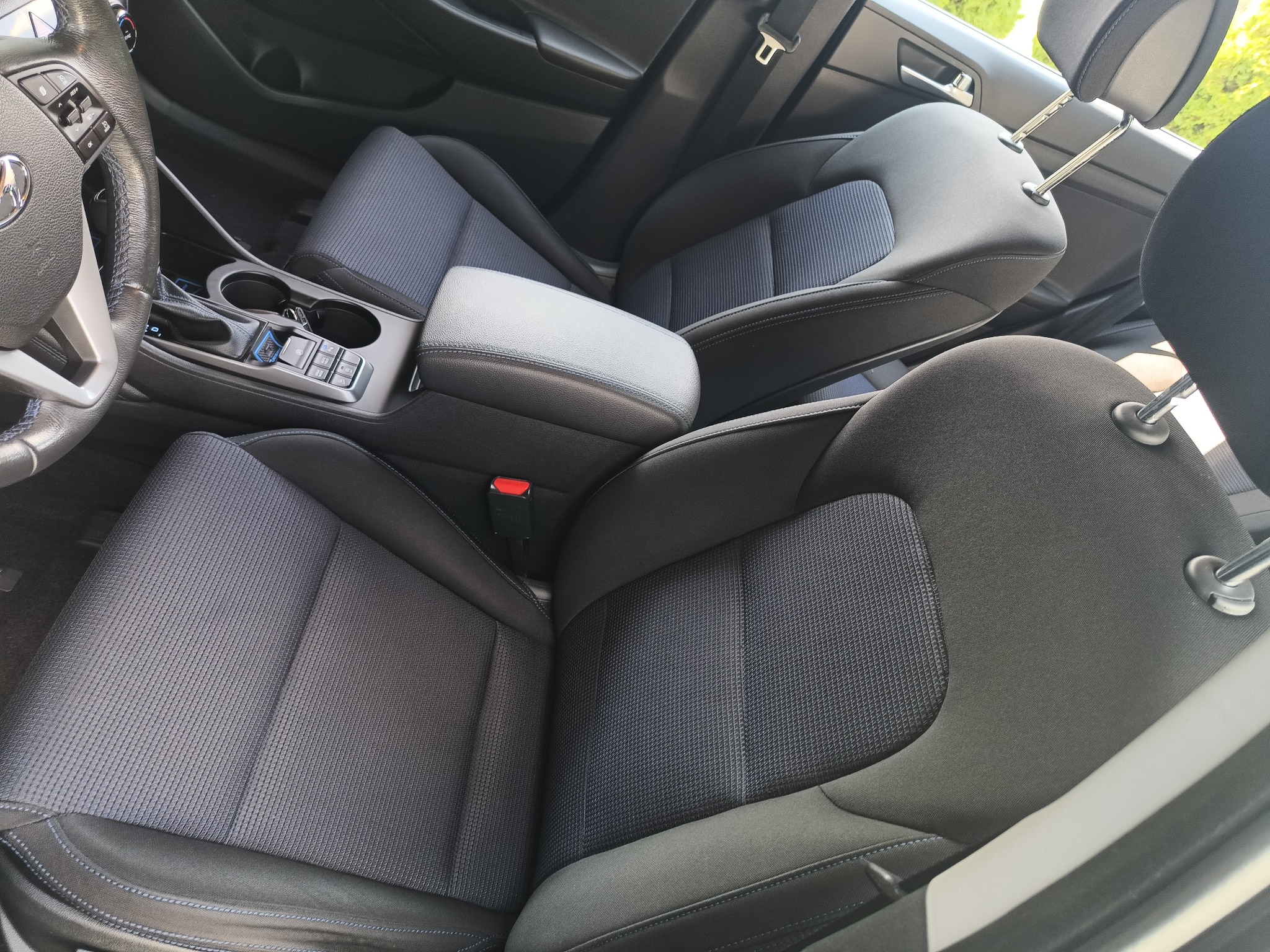 Wnętrze samochodu: czarne fotele z szarą tapicerką o drobnej strukturze, widoczny podłokietnik, kierownica i panel sterowania szybami oraz lusterkami. Ujęcie z góry, ukazujące detale wykończenia.