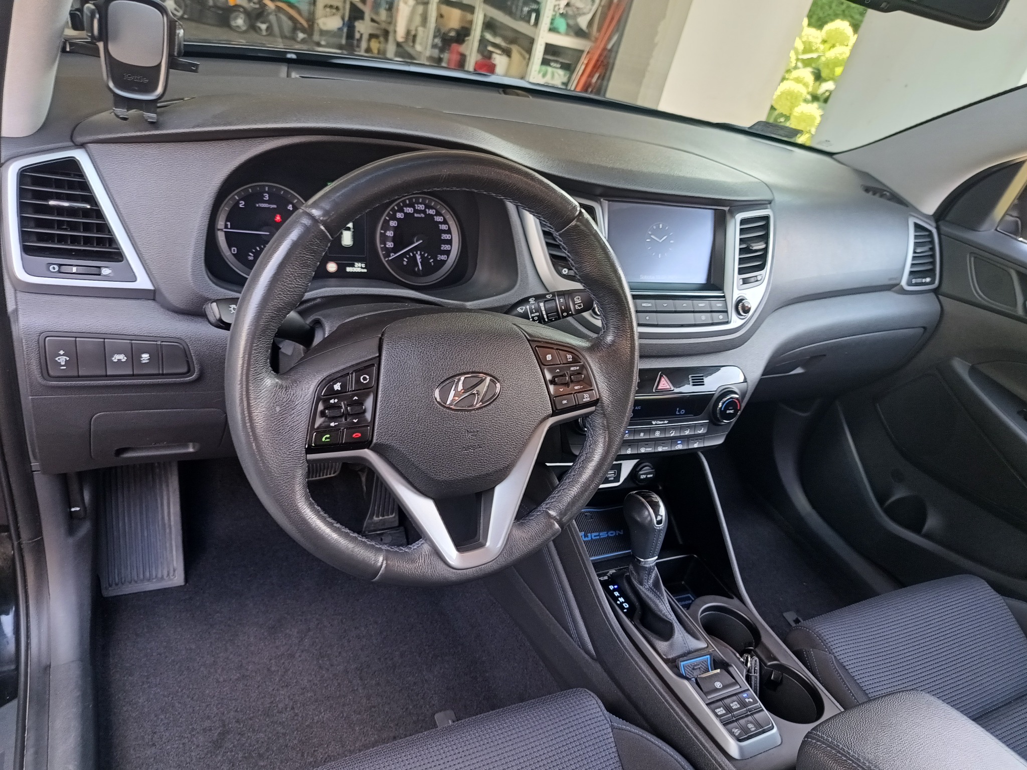 Wnętrze samochodu Hyundai Tucson, widok na deskę rozdzielczą, kierownicę, zegary i konsolę środkową. Ciemna tapicerka. Widoczny uchwyt na telefon na desce rozdzielczej.
