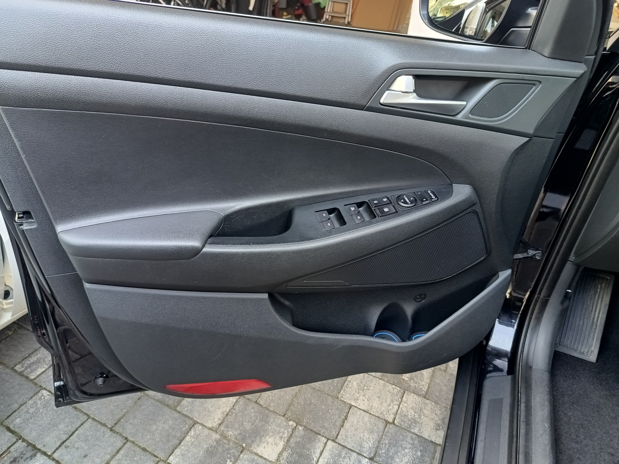 Wnętrze czarnego samochodu, widok na panel drzwi kierowcy z przyciskami sterowania szybami i lusterkami, schowkiem oraz głośnikiem.