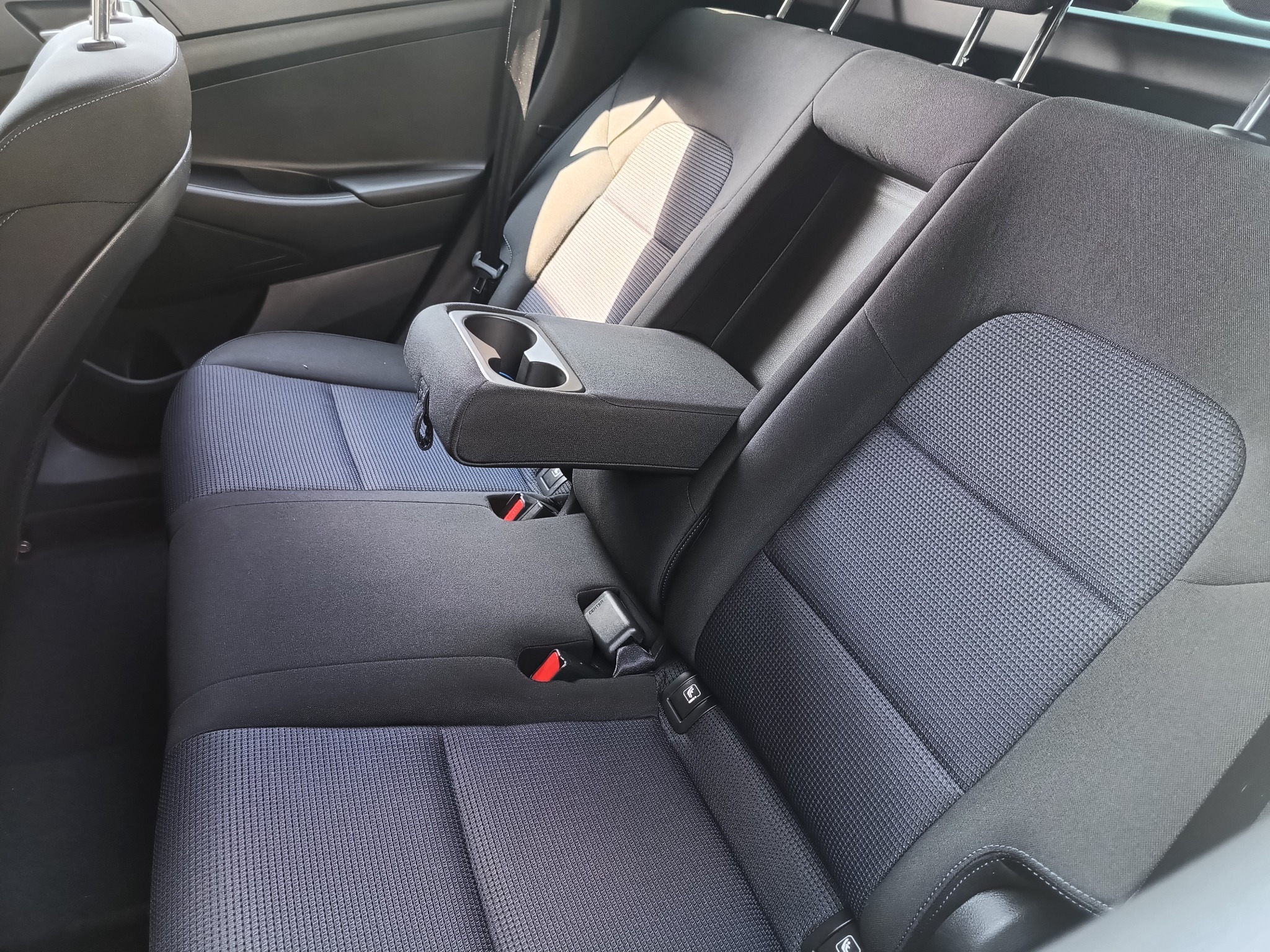 Wnętrze samochodu, tylna kanapa z podłokietnikiem i uchwytami na napoje, tapicerka w odcieniach szarości z delikatnym wzorem.