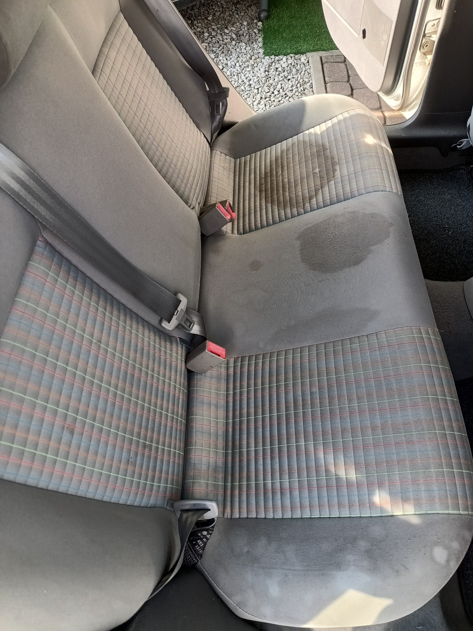 Brudna, szara tylna kanapa samochodowa z plamami i pasami bezpieczeństwa, widoczne elementy wnętrza pojazdu.