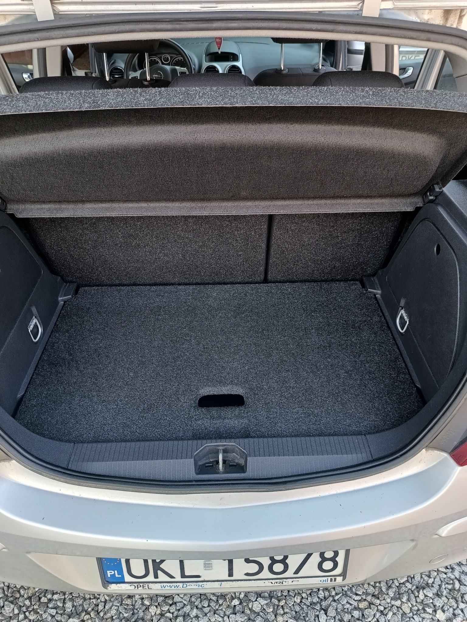 Otwarty bagażnik srebrnego samochodu Opel z widoczną szarą wykładziną, uchwytami mocującymi i tablicą rejestracyjną.