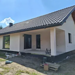 Parterowy dom z jasną, świeżo otynkowaną elewacją i ciemną dach&oacute;wką, widoczne ślady prac wykończeniowych wok&oacute;ł budynku, paleta i drabina na tarasie.