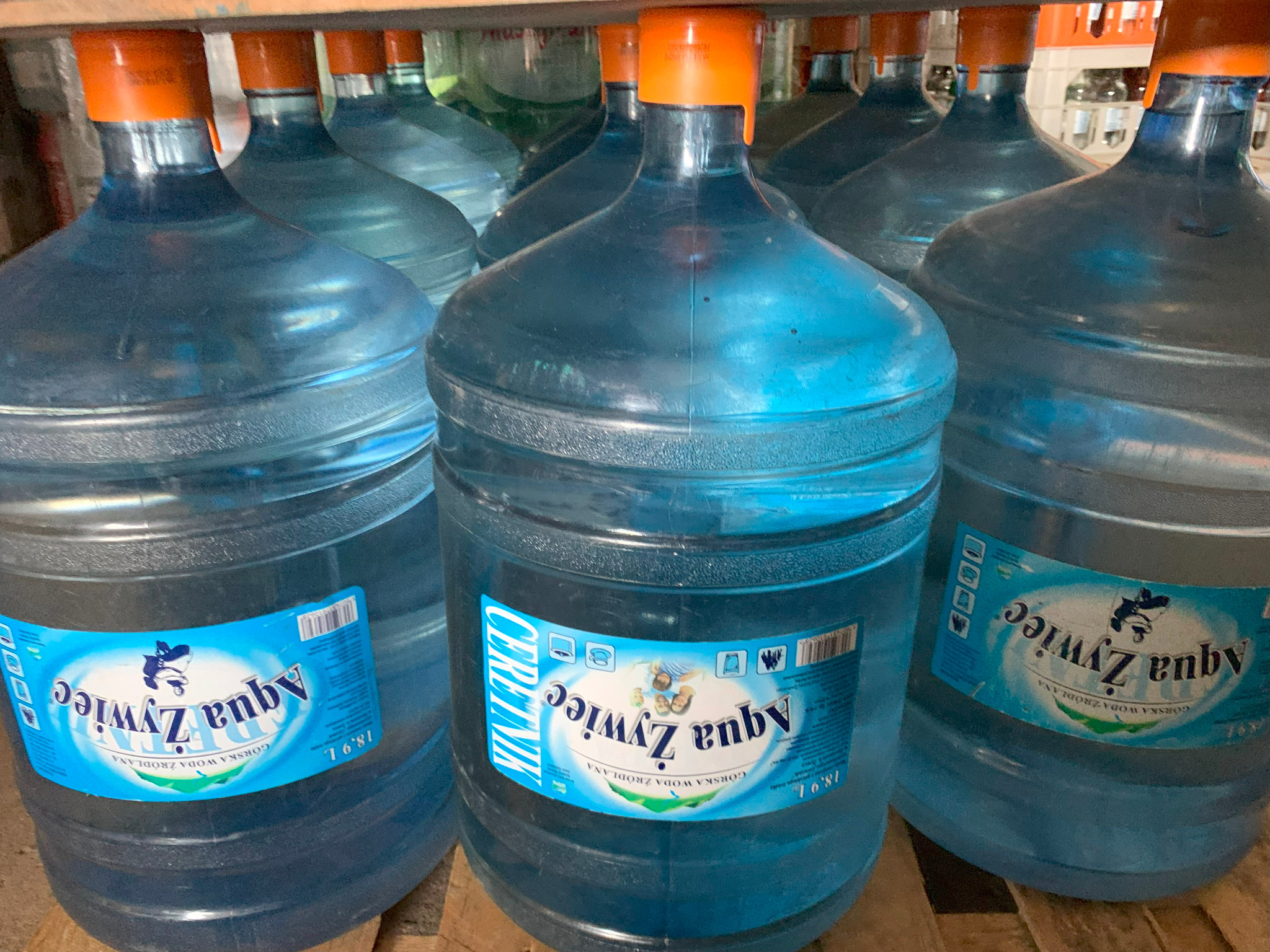 Baterie butli z wodą Aqua Żywiec 18.9L z pomarańczowymi nakrętkami, ustawione na drewnianej palecie, widoczne etykiety z nazwą i logo.