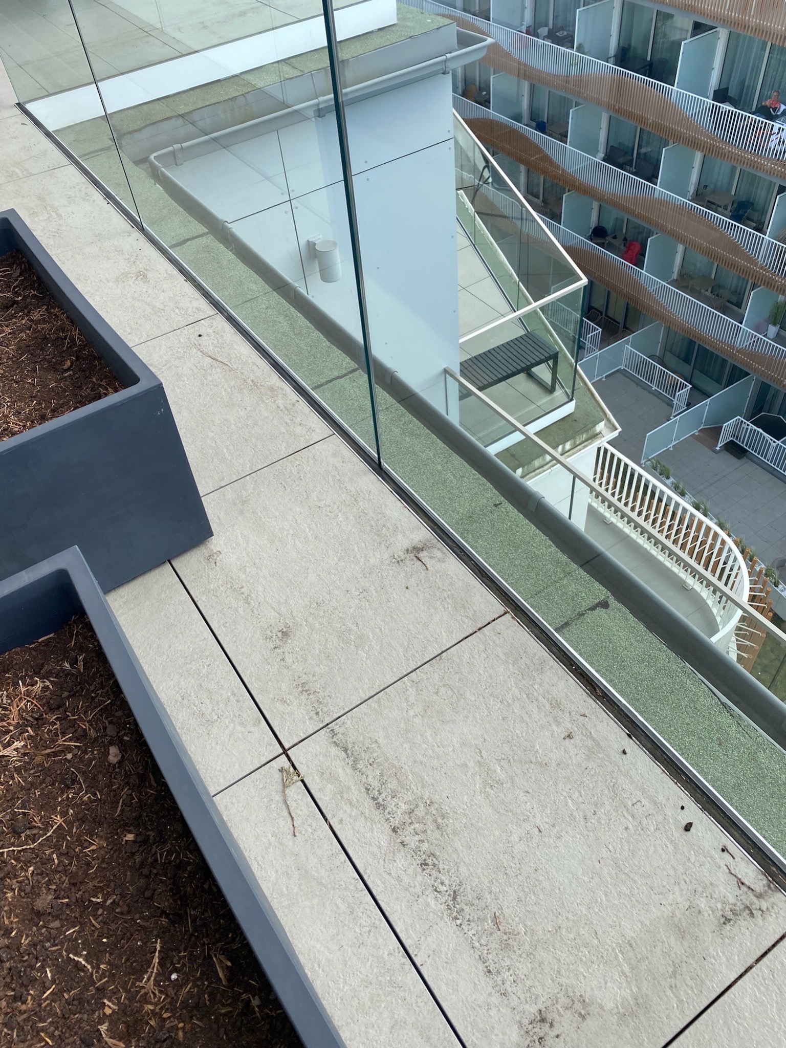 Widok z góry na balkon z szarymi płytkami, szklaną balustradą i donicą z ziemią, z widokiem na inne balkony w tle.