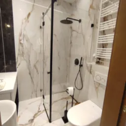 Nowoczesna łazienka z kabiną prysznicową typu walk-in, wykończona płytkami imitującymi marmur z wyraźnym, złotym użyleniem, czarną armaturą i białym wyposażeniem.