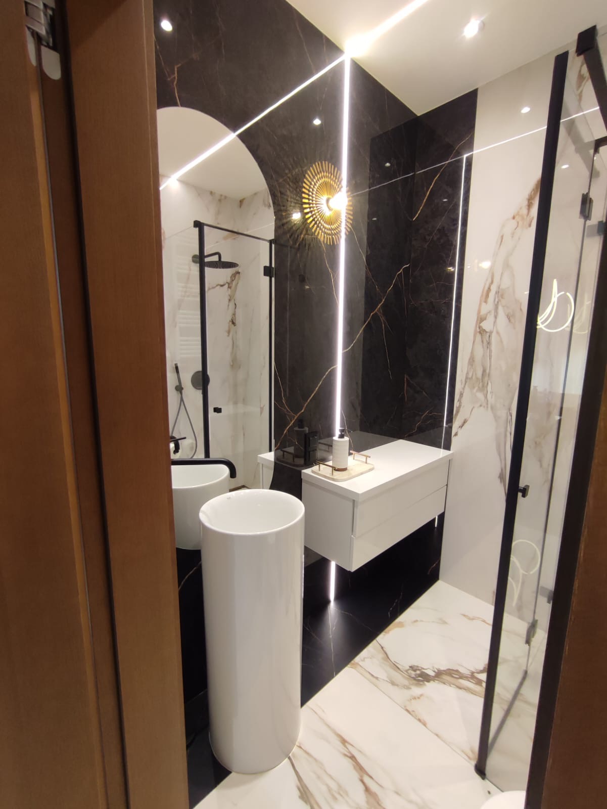 Nowoczesna łazienka z kabiną prysznicową typu walk-in, umywalką wolnostojącą i podświetlanym lustrem w kształcie słońca na ścianie z czarnego marmuru.