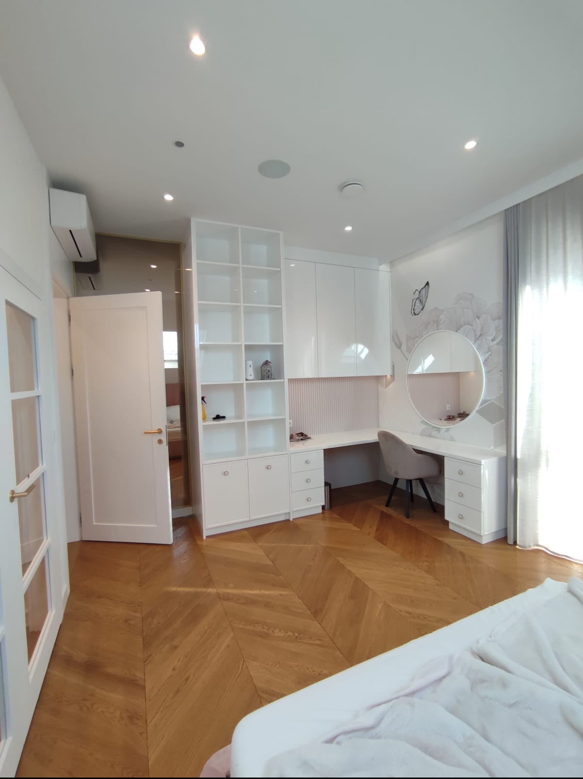 Jasne wnętrze sypialni z drewnianą podłogą w jodełkę, białymi meblami w zabudowie, toaletką z okrągłym lustrem i tapetą z motywem kwiatowym oraz motylem.