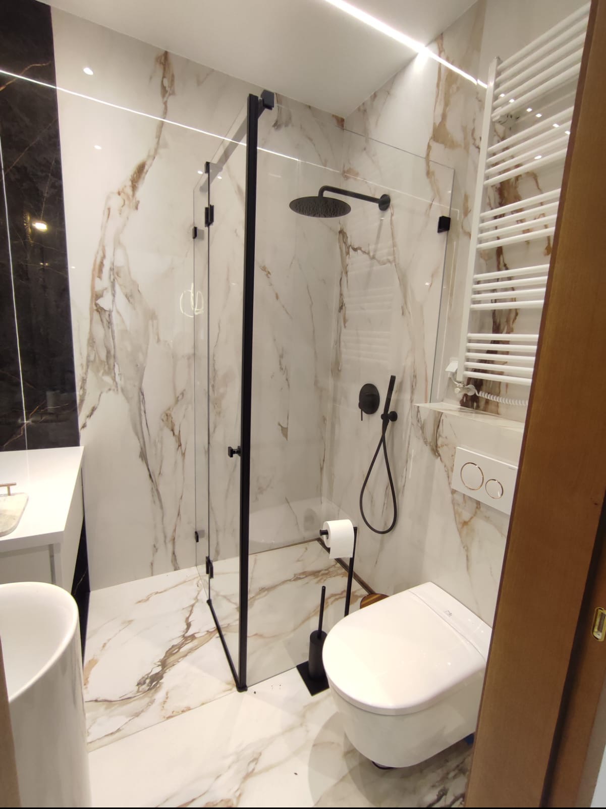 Nowoczesna łazienka z kabiną prysznicową typu walk-in, wykończona płytkami imitującymi marmur z wyraźnym, złotym użyleniem, czarną armaturą i białym wyposażeniem.