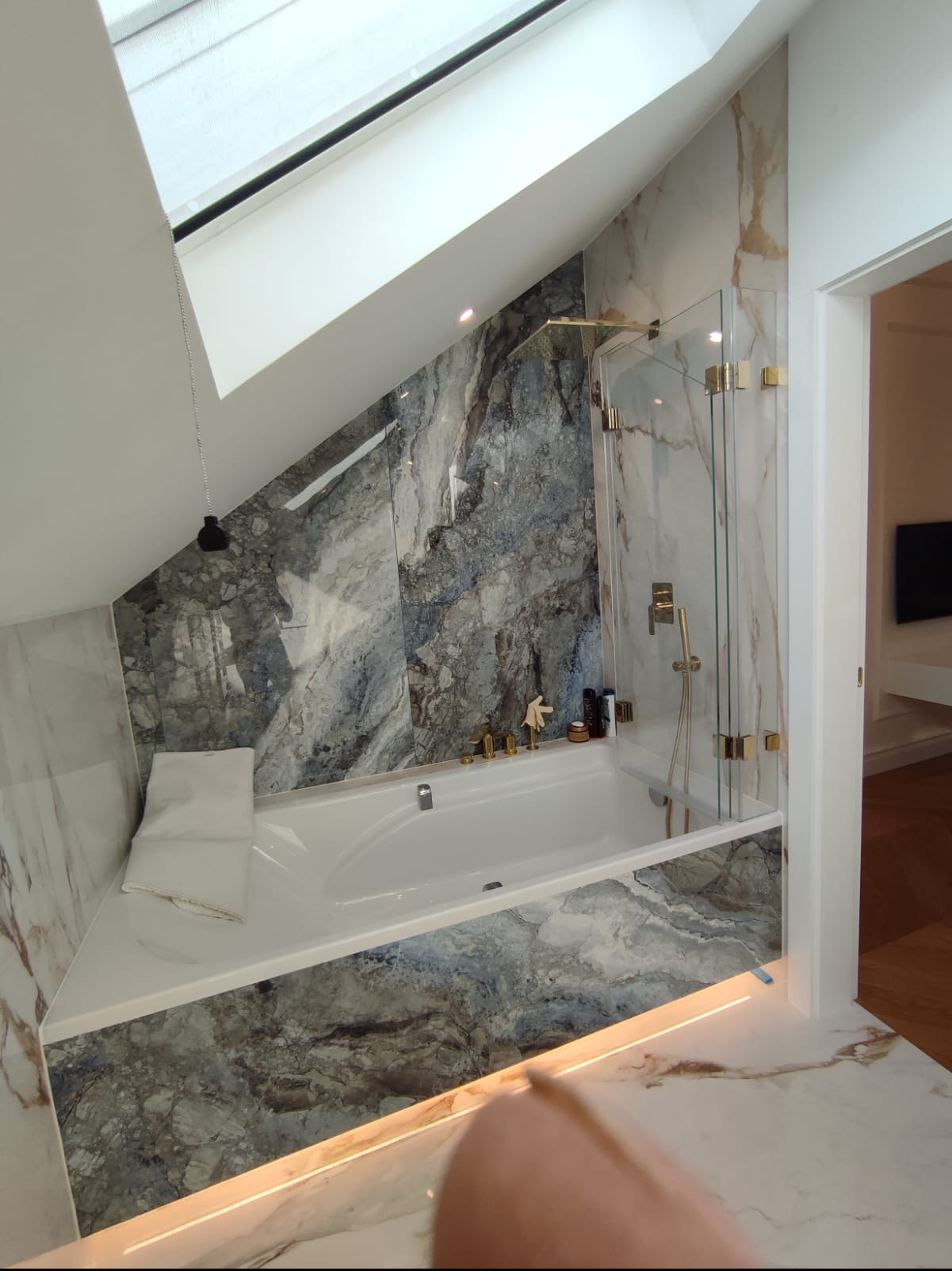 Nowoczesna łazienka na poddaszu z marmurowymi ścianami, białą wanną, szklanym parawanem i oknem dachowym z roletą.