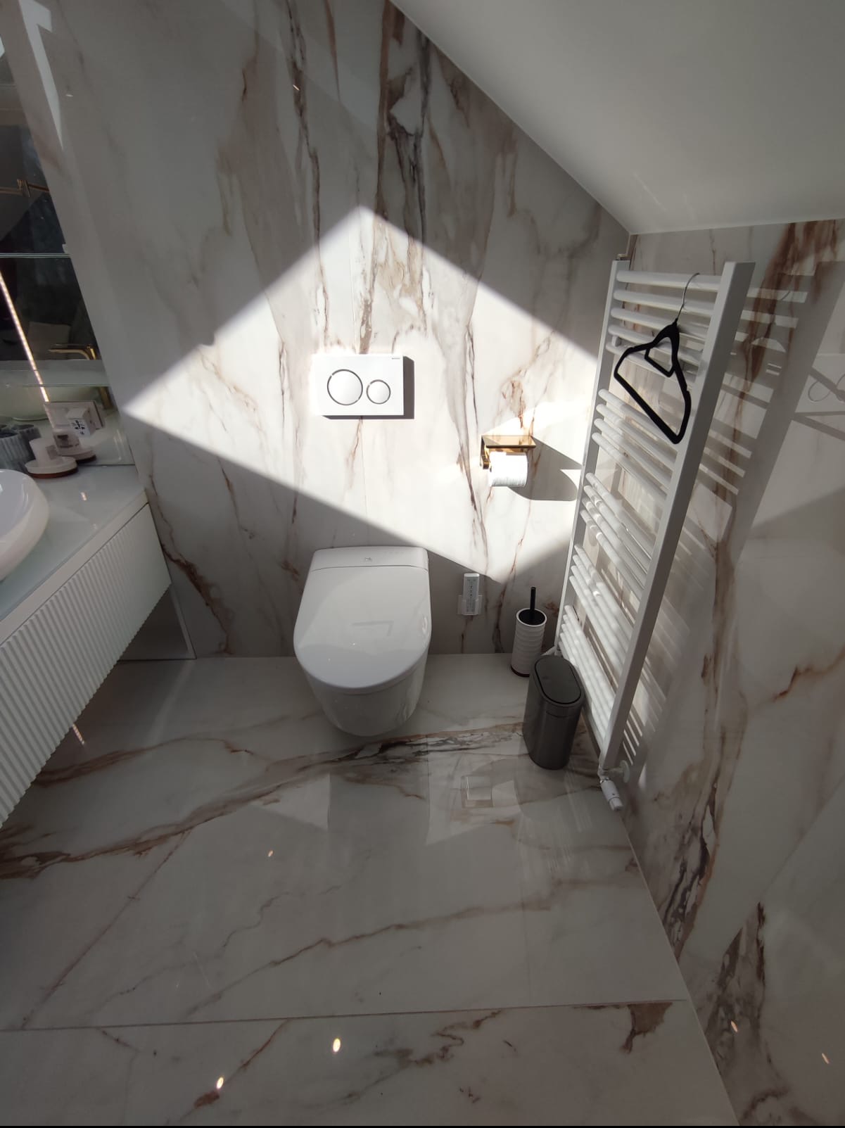 Nowoczesna toaleta z podwieszanym sedesem, marmurowymi płytkami na ścianach i podłodze, grzejnik drabinkowy z wieszakiem, oświetlona naturalnym światłem.