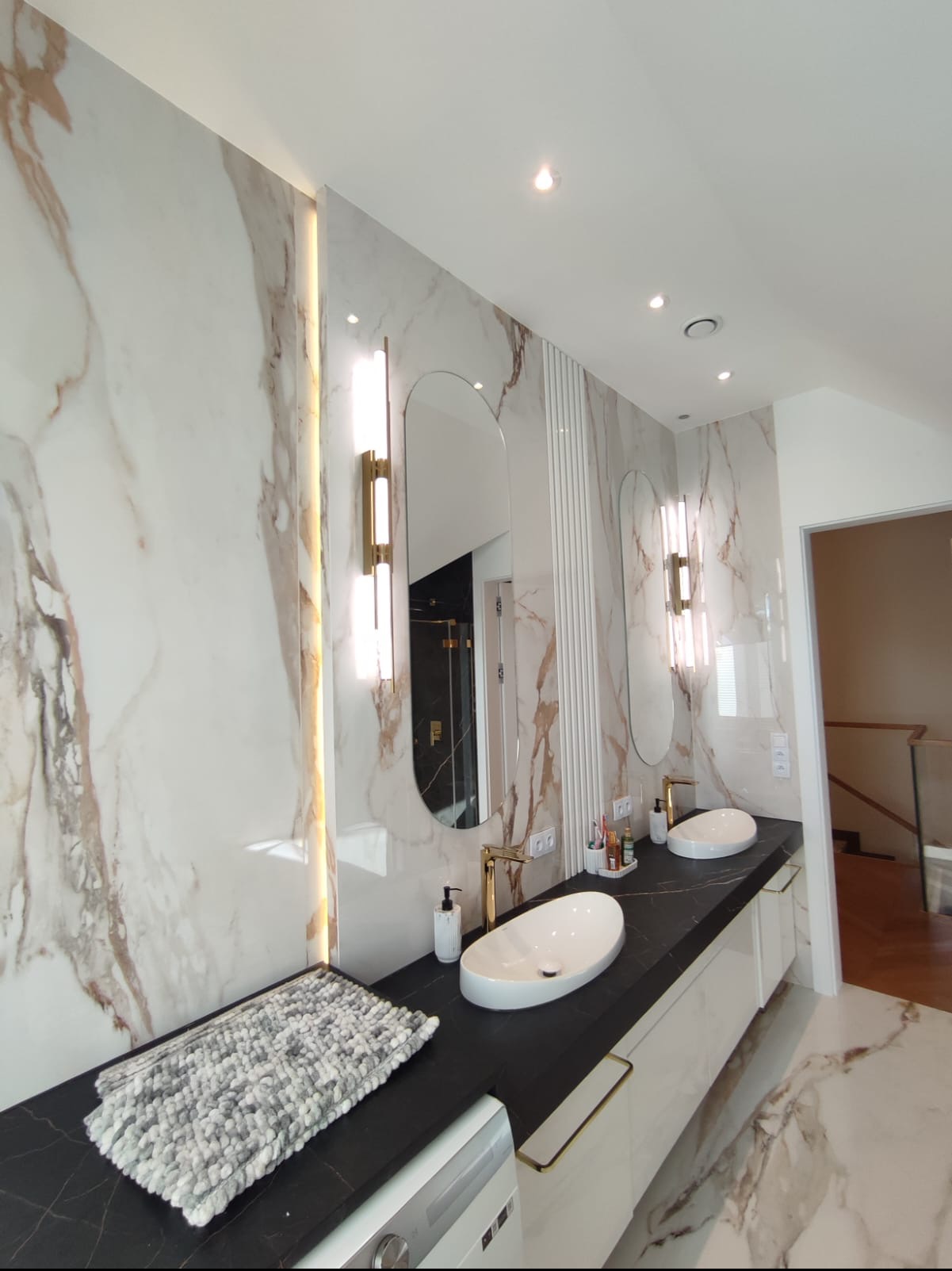 Nowoczesna łazienka z marmurowymi płytkami, owalnymi lustrami podświetlonymi lampami, czarnym blatem i białymi umywalkami nablatowymi, uzupełniona szarą matą i złotymi detalami.