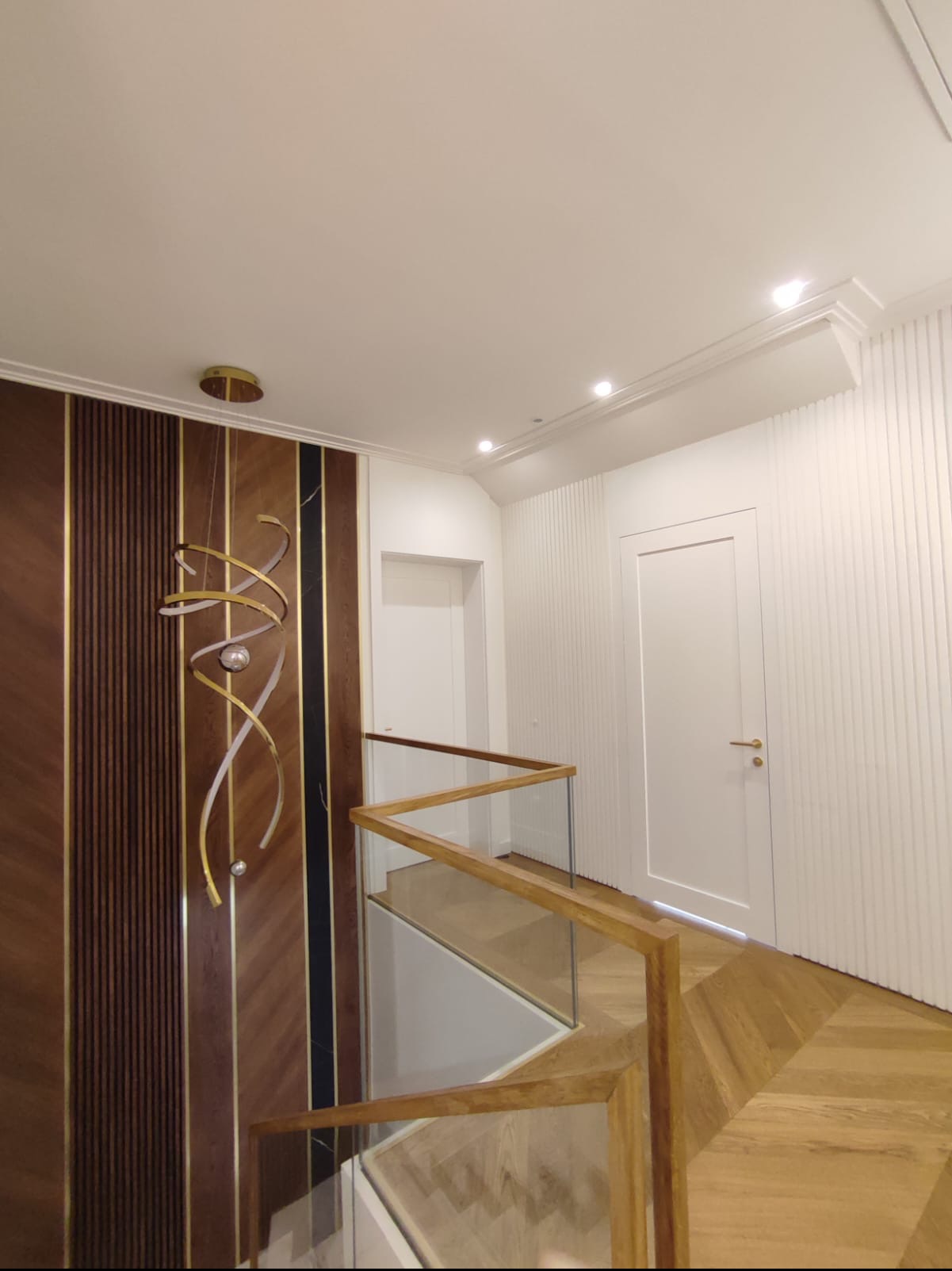 Nowoczesne wnętrze domu z drewnianą boazerią, szklaną balustradą schodów i designerską lampą wiszącą z elementami złota.