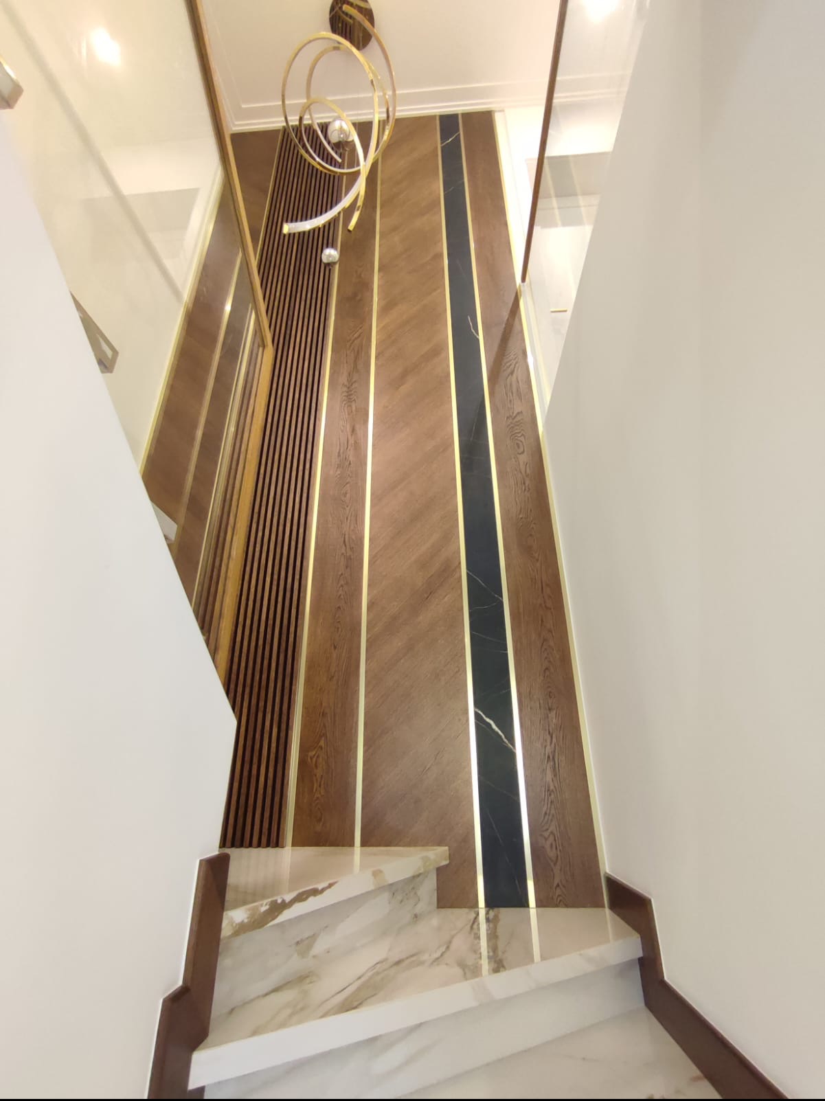 Nowoczesna klatka schodowa z marmurowymi stopniami, drewnianymi panelami ściennymi z akcentami w kolorze złota i designerską lampą wiszącą.